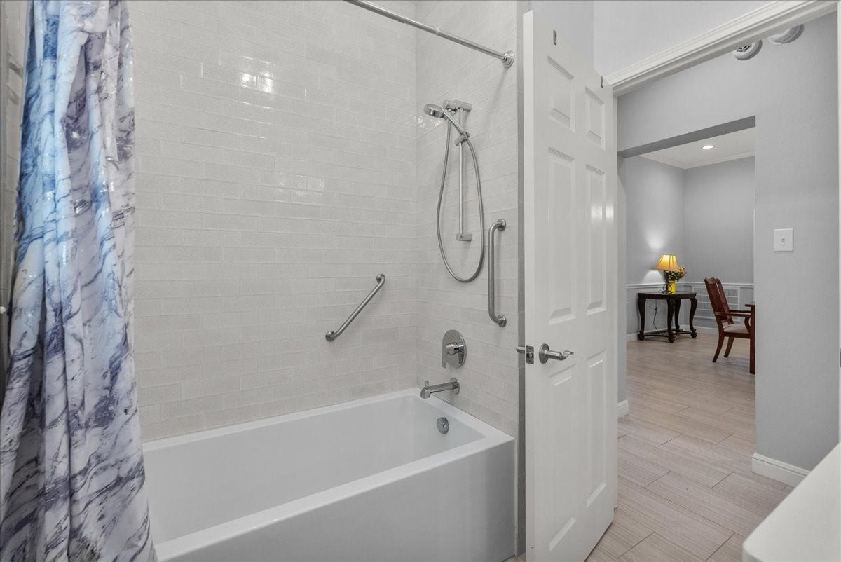 This bathroom features a modern design with white subway tiles, a shower-tub combo, and a sleek marble-patterned shower curtain. It includes a safety grab bar and opens to a well-lit adjoining room with wooden furniture.
