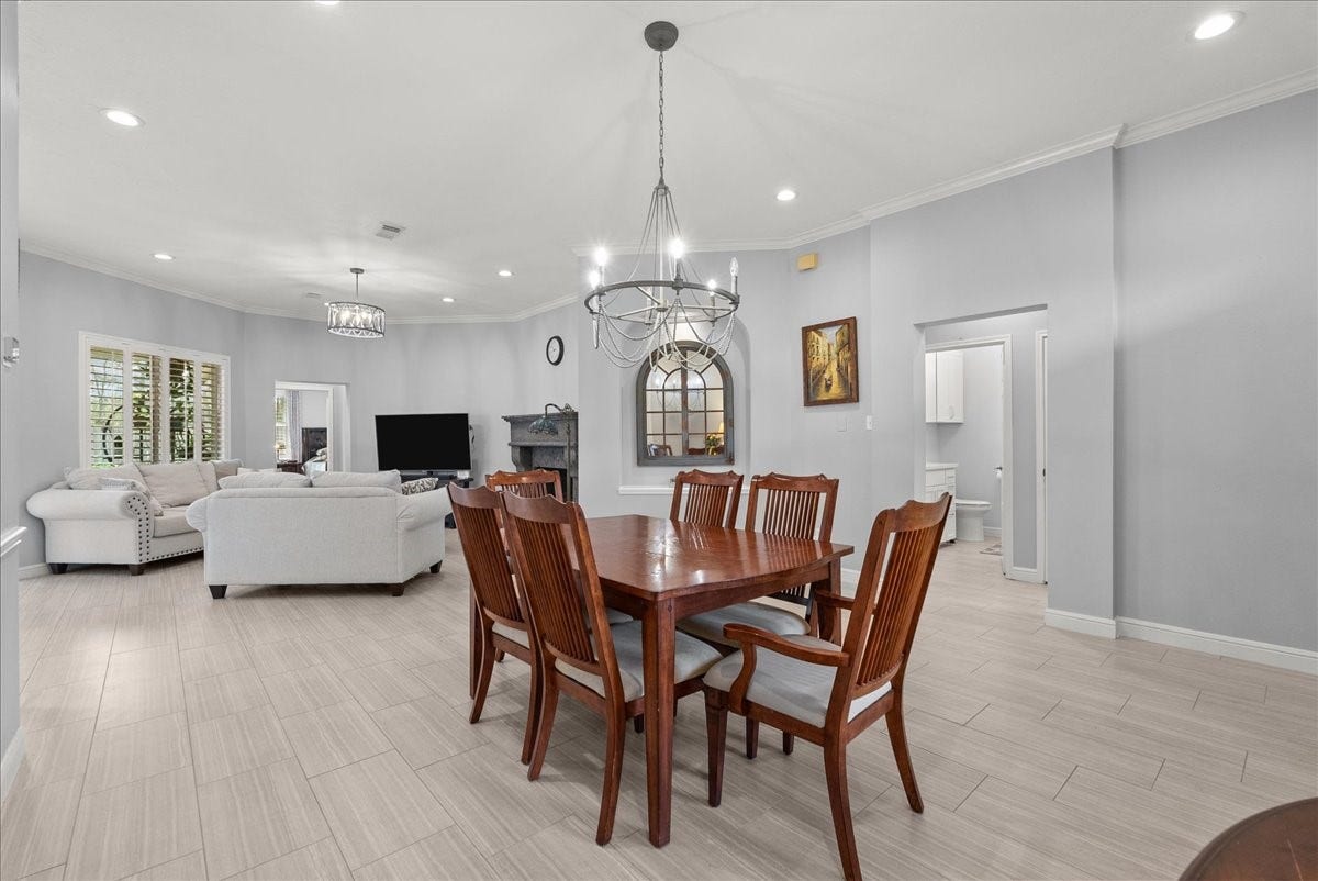 This photo features a spacious open-concept living and dining area with neutral tones and tile flooring. The dining space has a wooden table set under a modern chandelier, while the living area is furnished with light-colored sofas and a wall-mounted TV. Large windows with shutters provide natural light, and there's a cozy fireplace for added warmth.
