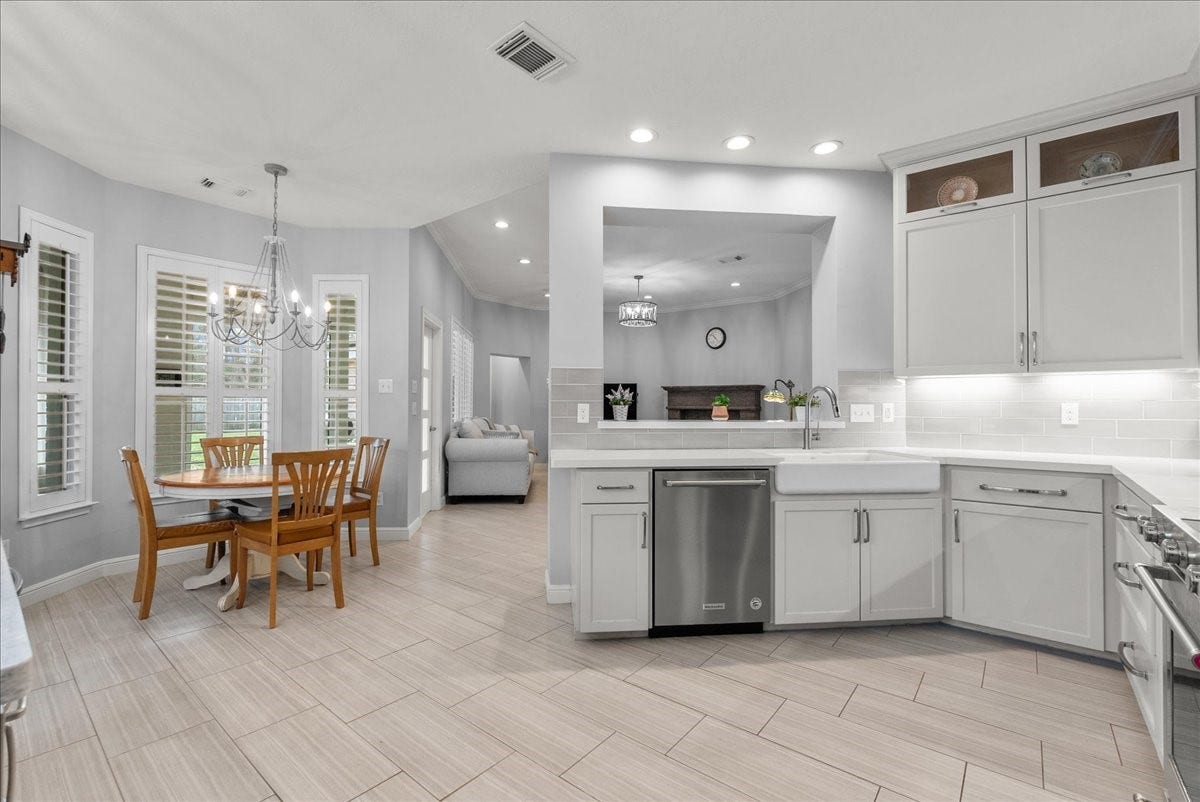 This photo showcases a modern, open-concept kitchen and dining area. The kitchen features light gray cabinets, stainless steel appliances, and a farmhouse sink. The dining area has a wooden table set beneath a stylish chandelier, and large windows with shutters bring in natural light. The neutral tones and recessed lighting create a bright and inviting atmosphere.