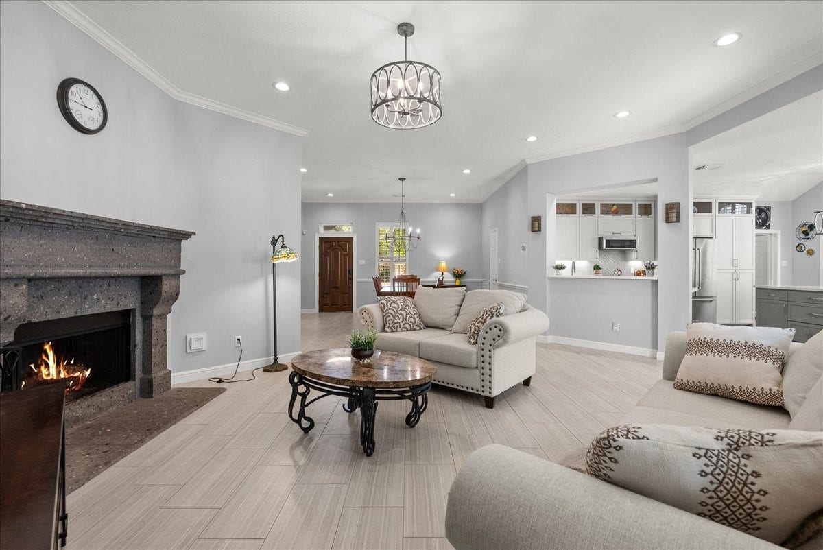 Spacious living room with a cozy fireplace, light gray walls, and tile flooring. The open layout connects to a modern kitchen with a breakfast bar, creating a welcoming and functional space.