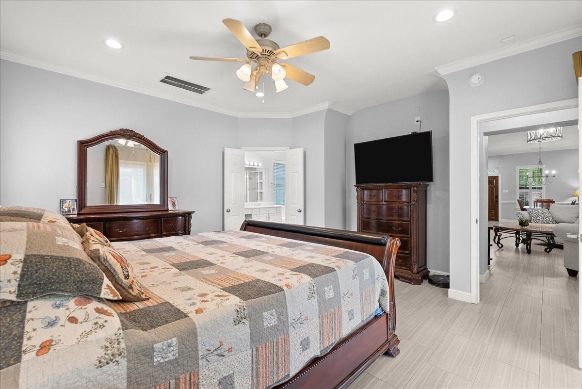 This bedroom features a comfortable, spacious layout with a large bed and elegant wooden furniture, including a dresser and a chest with a mounted TV. Soft gray walls and light flooring create a serene atmosphere, while a ceiling fan adds comfort. The room opens up to a bright bathroom and a cozy living area, enhancing its functionality and flow.