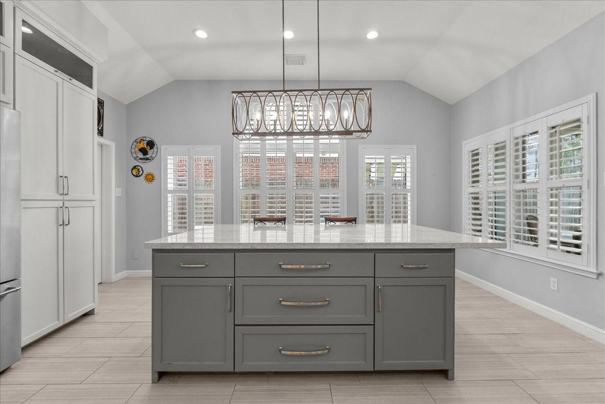 Modern kitchen with a large island and marble countertop. Features sleek cabinetry, stainless steel appliances, and ample natural light through plantation shutters. Ideal for cooking and entertaining.