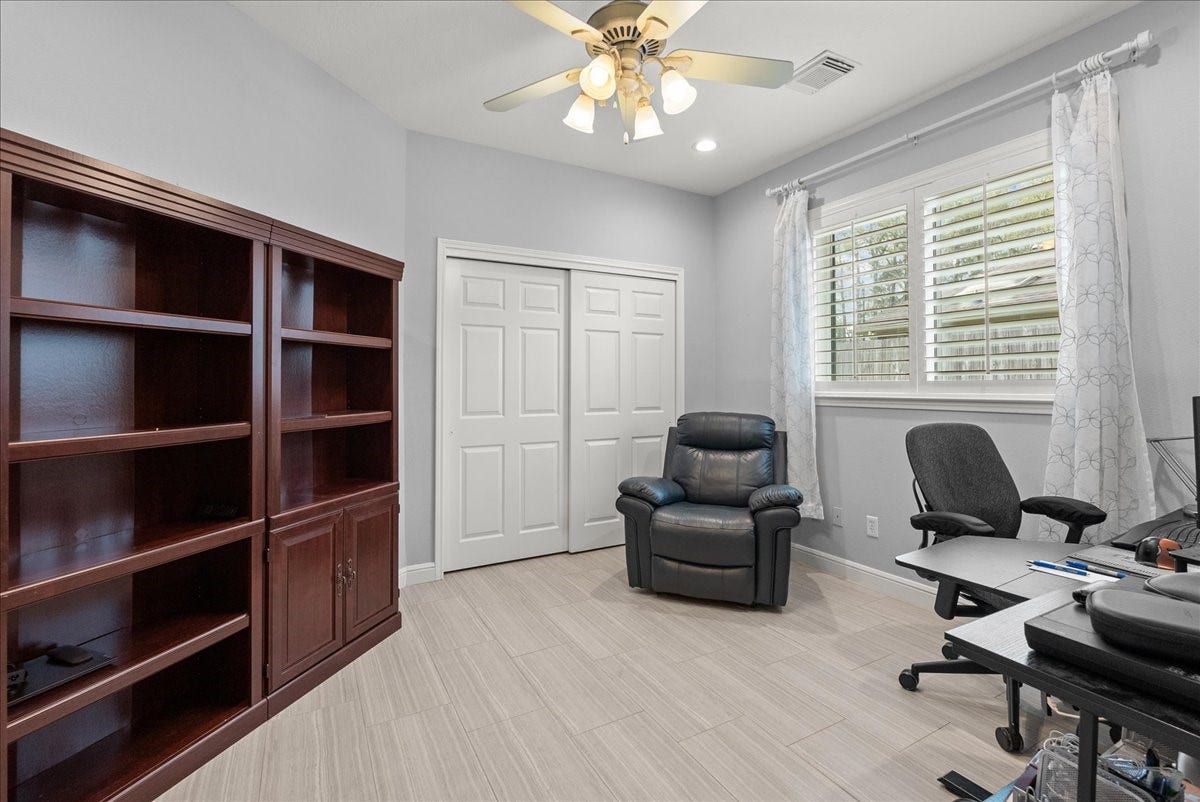 This room offers a cozy workspace with a comfortable chair, desk, and ample natural light from a large window with blinds. It features built-in shelving for storage and a ceiling fan for comfort. Ideal for a home office or study area.