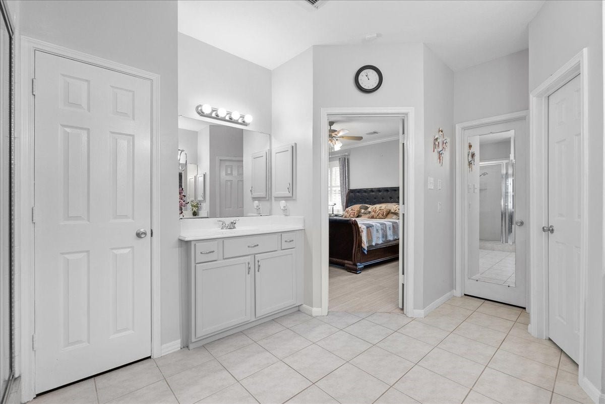 This photo shows a bright, clean bathroom with a double vanity and ample storage. It features a large mirror, modern lighting, and tile flooring. The room connects to a bedroom with a glimpse of a bed and ceiling fan, and there are multiple doors leading to other spaces, including a closet with a mirrored door.