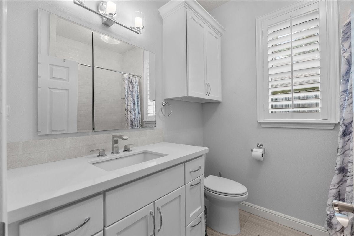 This bathroom features a clean, modern design with white cabinetry and countertops. It includes a large mirror, contemporary fixtures, and a window with plantation shutters, offering a bright and airy feel.