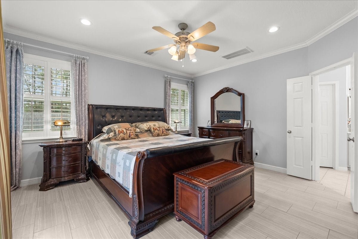 This is a spacious and elegant bedroom featuring a large bed with a tufted headboard, wooden furniture, and light-colored walls. It has ample natural light from the windows with plantation shutters, and a ceiling fan for comfort. The room connects to an ensuite bathroom, adding convenience.
