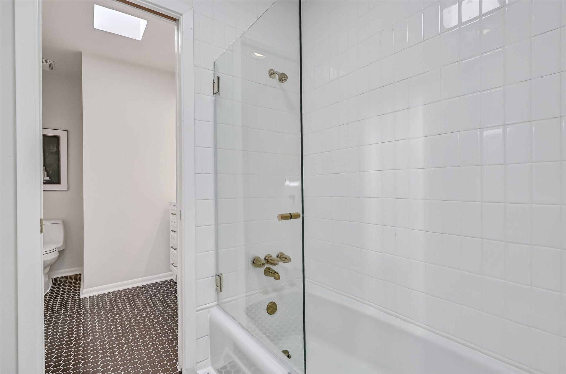 300 Courtlandt Place, Friendswood, TX 77546 The shared bathing area features a tub/shower combination with white tiled walls, a frameless glass panel & gold-tone plumbing fixtures. Black hexagonal tile flooring continues throughout, while a separate toilet compartment and individual vanity areas provide added functionality for both bedrooms.