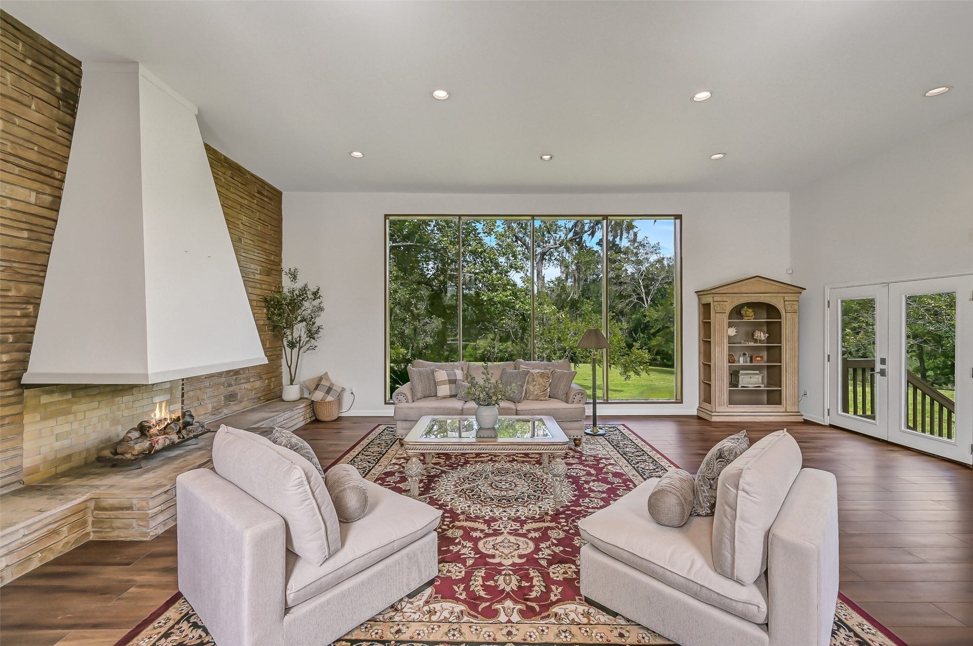 300 Courtlandt Place, Friendswood, TX 77546 An impressive wall of picture windows brings the outdoors into the spacious living room, offering sweeping views across the backyard and mature trees. The room's generous proportions provide flexibility for multiple seating arrangements, while the distinctive fireplace creates a striking focal point.