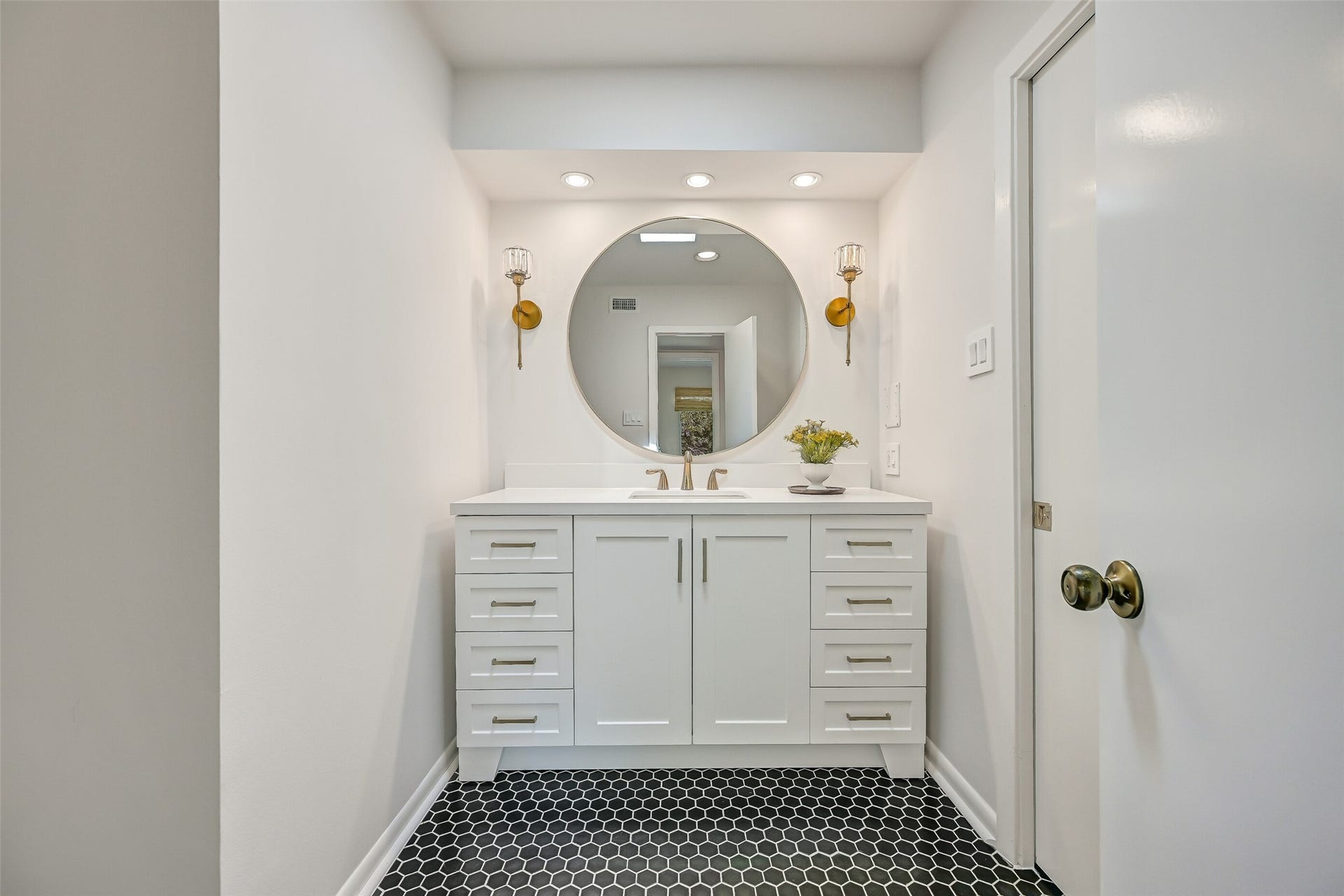 300 Courtlandt Place, Friendswood, TX 77546 One of two separate vanity areas in the Jack & Jill bathroom, this updated space features a white vanity with generous drawer storage, contemporary hardware, quartz-look countertop & round mirror flanked by decorative gold sconces. Black hexagonal tile flooring adds a striking contrast.