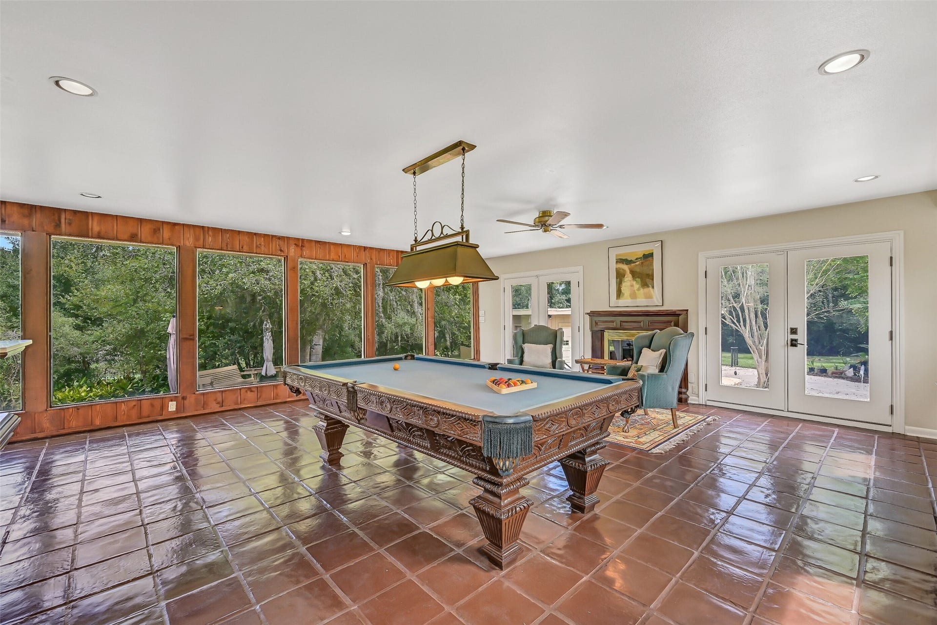300 Courtlandt Place, Friendswood, TX 77546 A different perspective showcases the gameroom's beautiful backyard views, with oversized windows framed by rich wood paneling. The pool table sits beneath a decorative hanging light, while a cozy seating area beside the second gas-log fireplace creates another inviting place to gather.