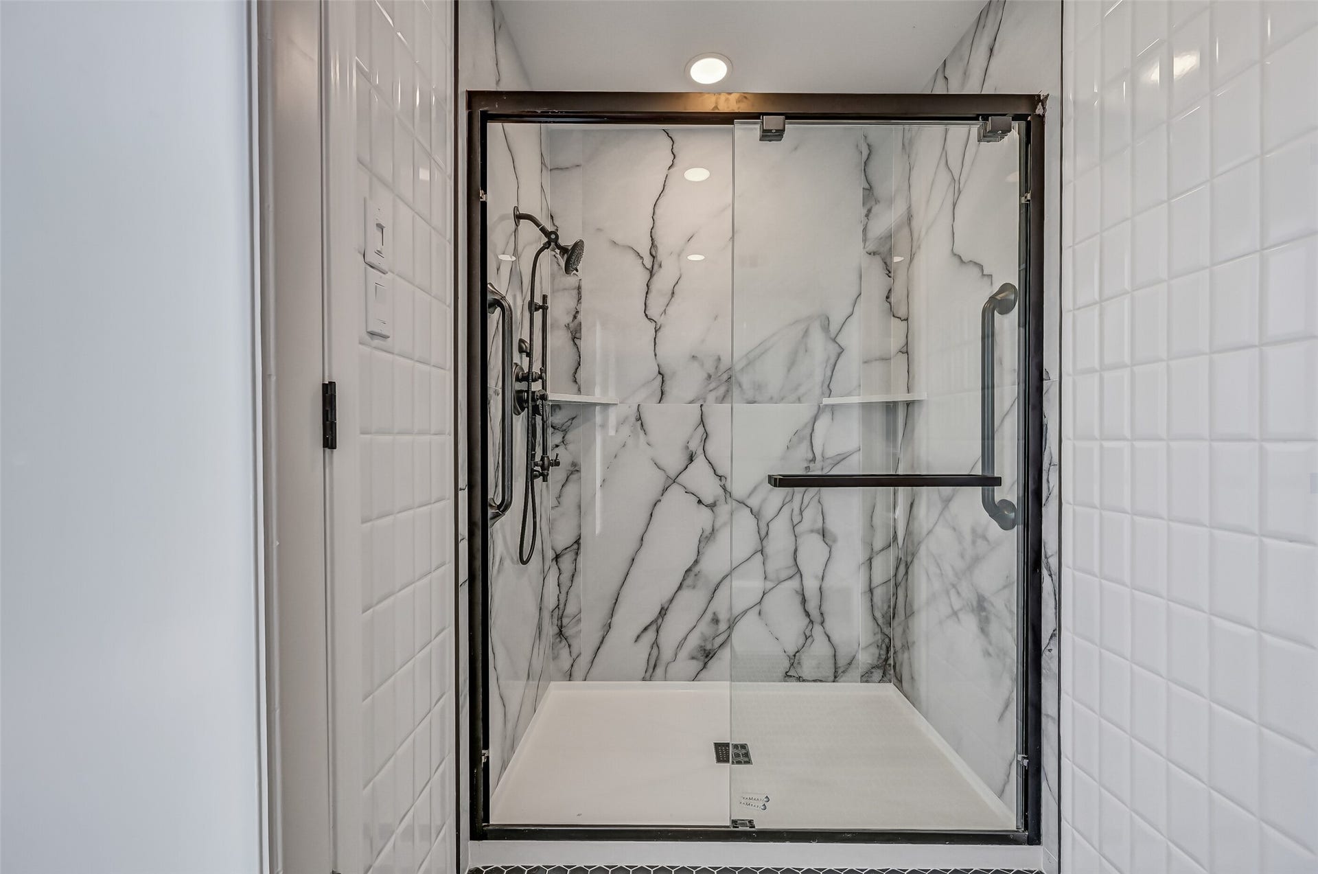 300 Courtlandt Place, Friendswood, TX 77546 The updated primary shower features dramatic marble-look wall surrounds, a sliding glass enclosure with dark metal framing, built-in corner shelves & a handheld shower fixture. Its generous dimensions provide a comfortable bathing space, complemented by the bathroom's black hexagonal tile flooring.