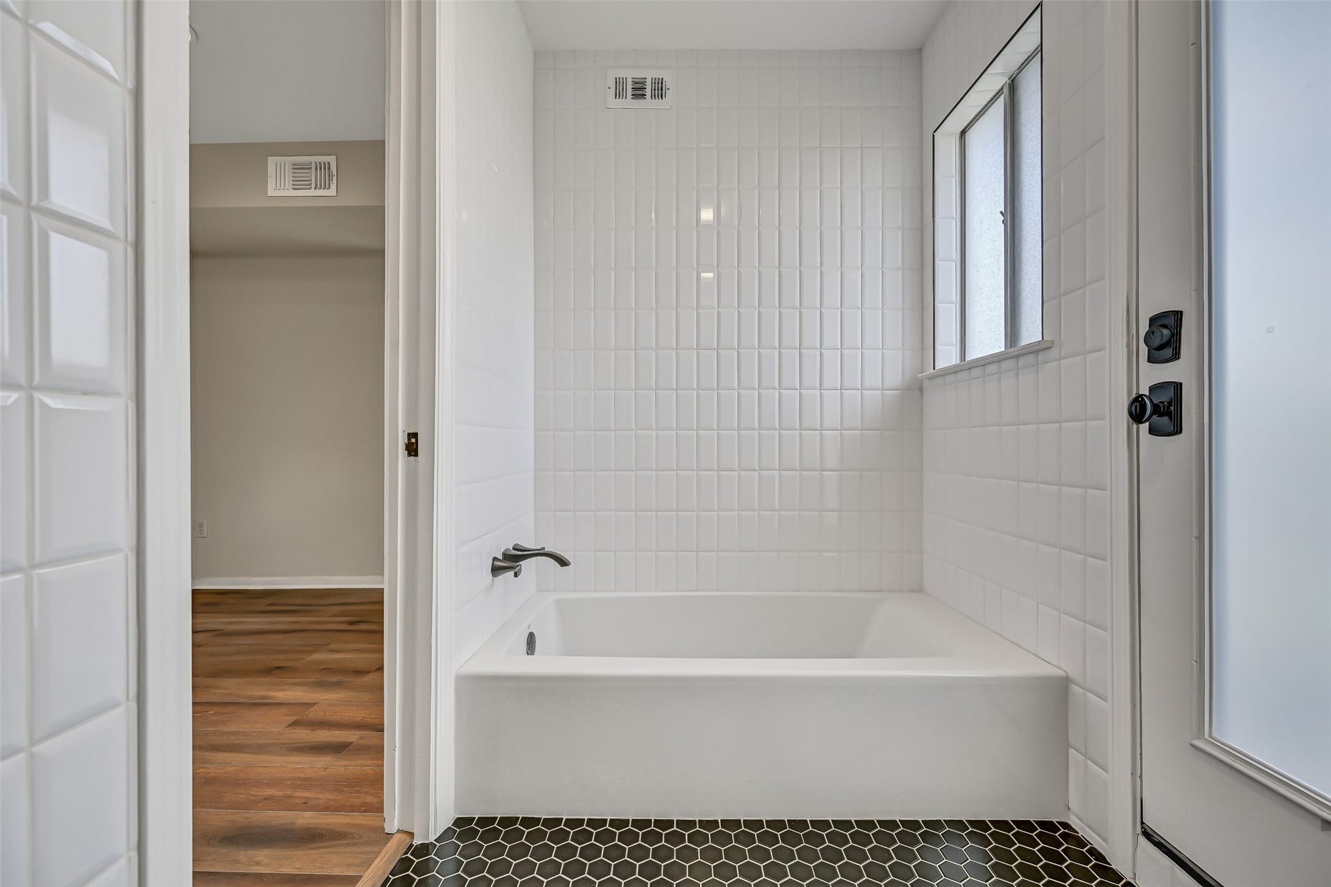 300 Courtlandt Place, Friendswood, TX 77546 A recently replaced soaking tub sits within a white tiled alcove in the primary bathroom, offering a separate bathing option alongside the updated shower. Black hexagonal tile flooring creates a contrasting finish, while a nearby exterior door provides access to the adjacent roof area.