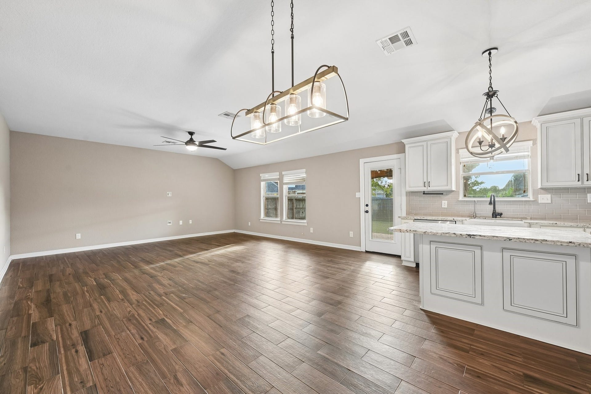 5012 Bayou Dr, Dickinson, TX 77539 Spacious open-concept living area with elegant wood flooring, modern light fixtures, and a bright kitchen featuring granite countertops and white cabinetry. Natural light floods through large windows and a glass patio door.