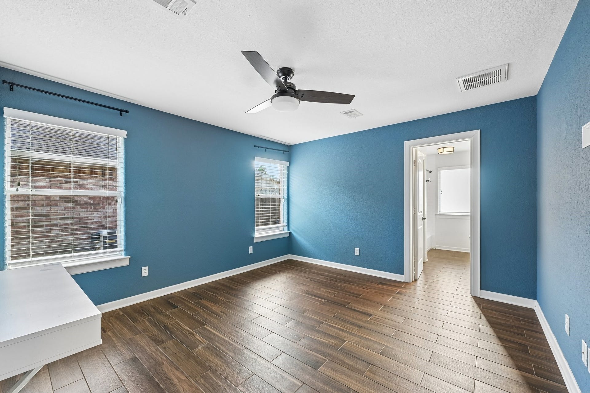 5012 Bayou Dr, Dickinson, TX 77539 Bright, spacious room featuring elegant wood-look flooring, soothing blue walls, and a modern ceiling fan. Abundant natural light flows through large windows, leading to an adjoining bathroom.