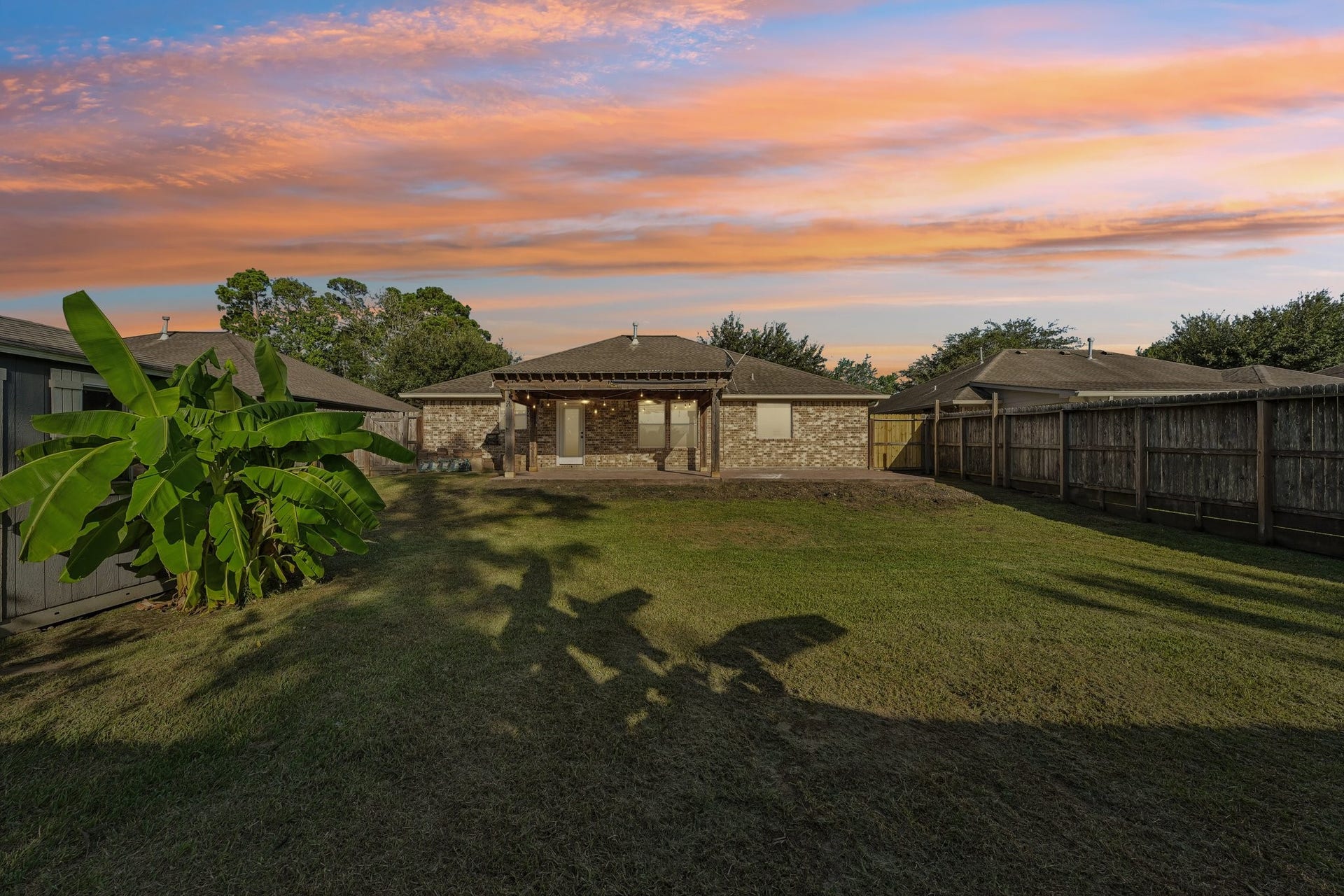 5012 Bayou Dr, Dickinson, TX 77539 Spacious backyard with well-maintained lawn, privacy fence, and a charming brick exterior under a stunning sunset sky.