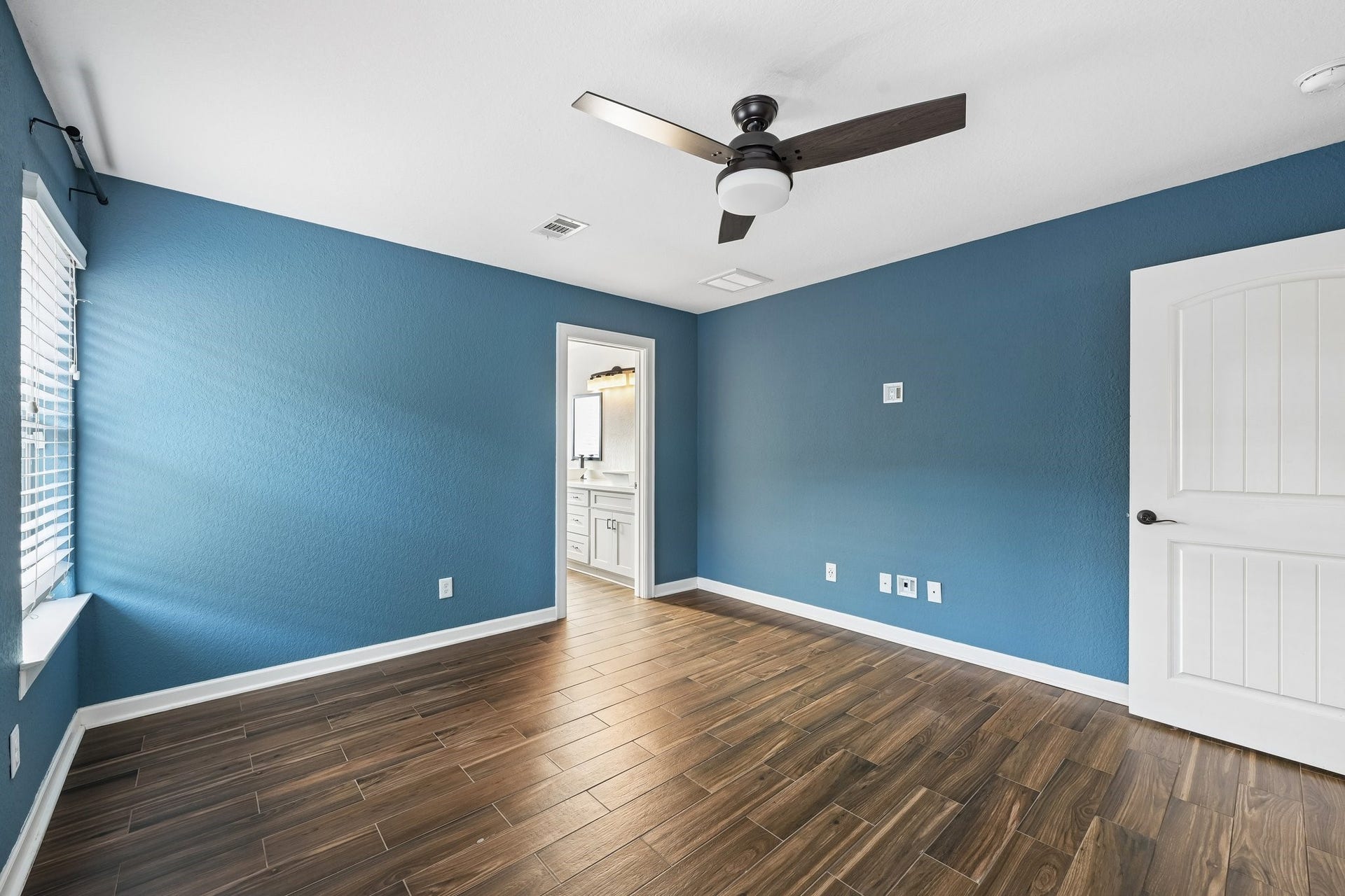 5012 Bayou Dr, Dickinson, TX 77539 Bright room with rich wood flooring, calming blue walls, and a modern ceiling fan. Natural light streams through large windows, leading to an elegant ensuite bathroom.