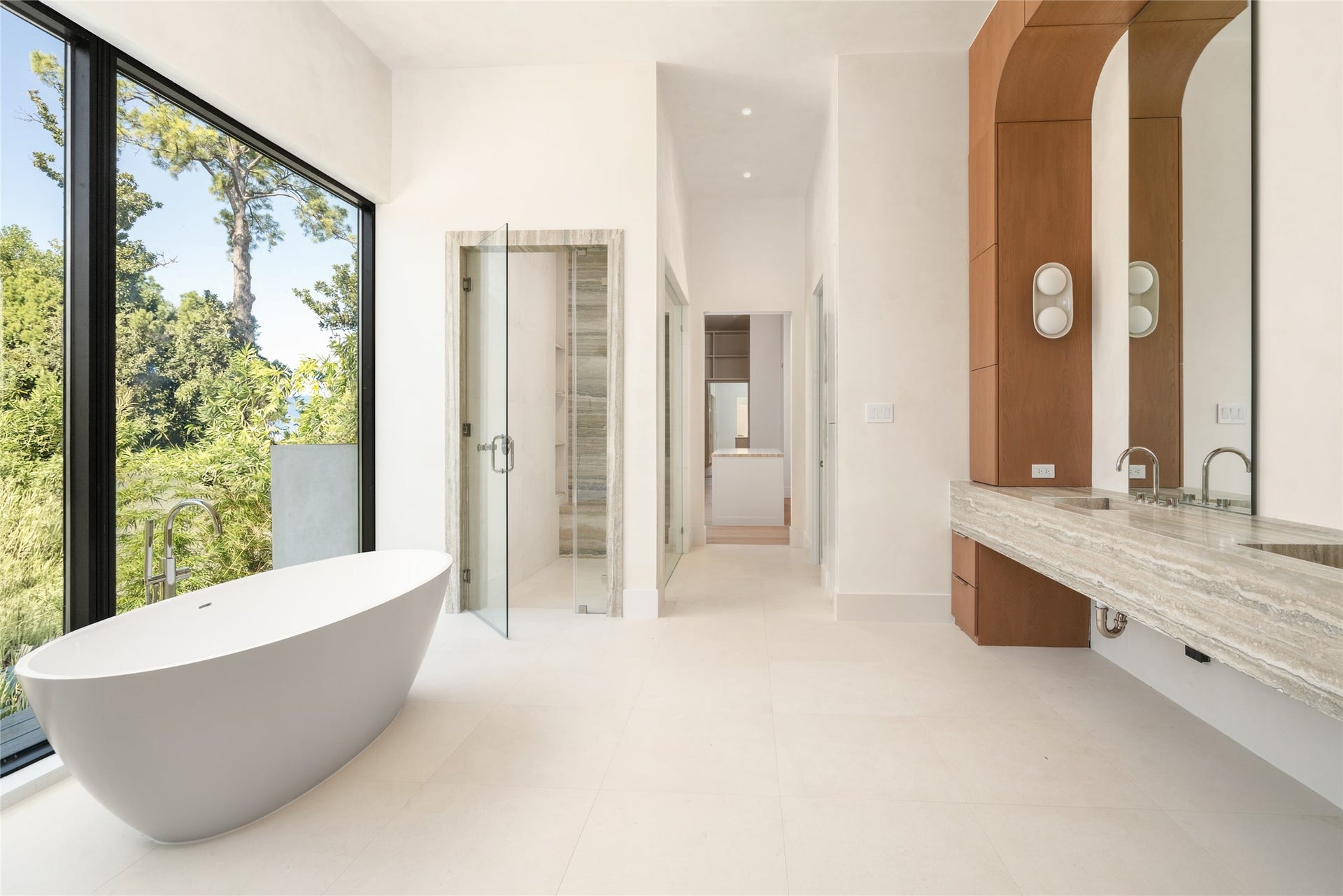 11 Tiel Way, Houston, TX 77019 The primary bath pairs a sculptural freestanding soaking tub with a floating double vanity, Silver Travertine finishes, and an oversized picture window overlooking the greenery, creating a serene, spa-inspired setting.