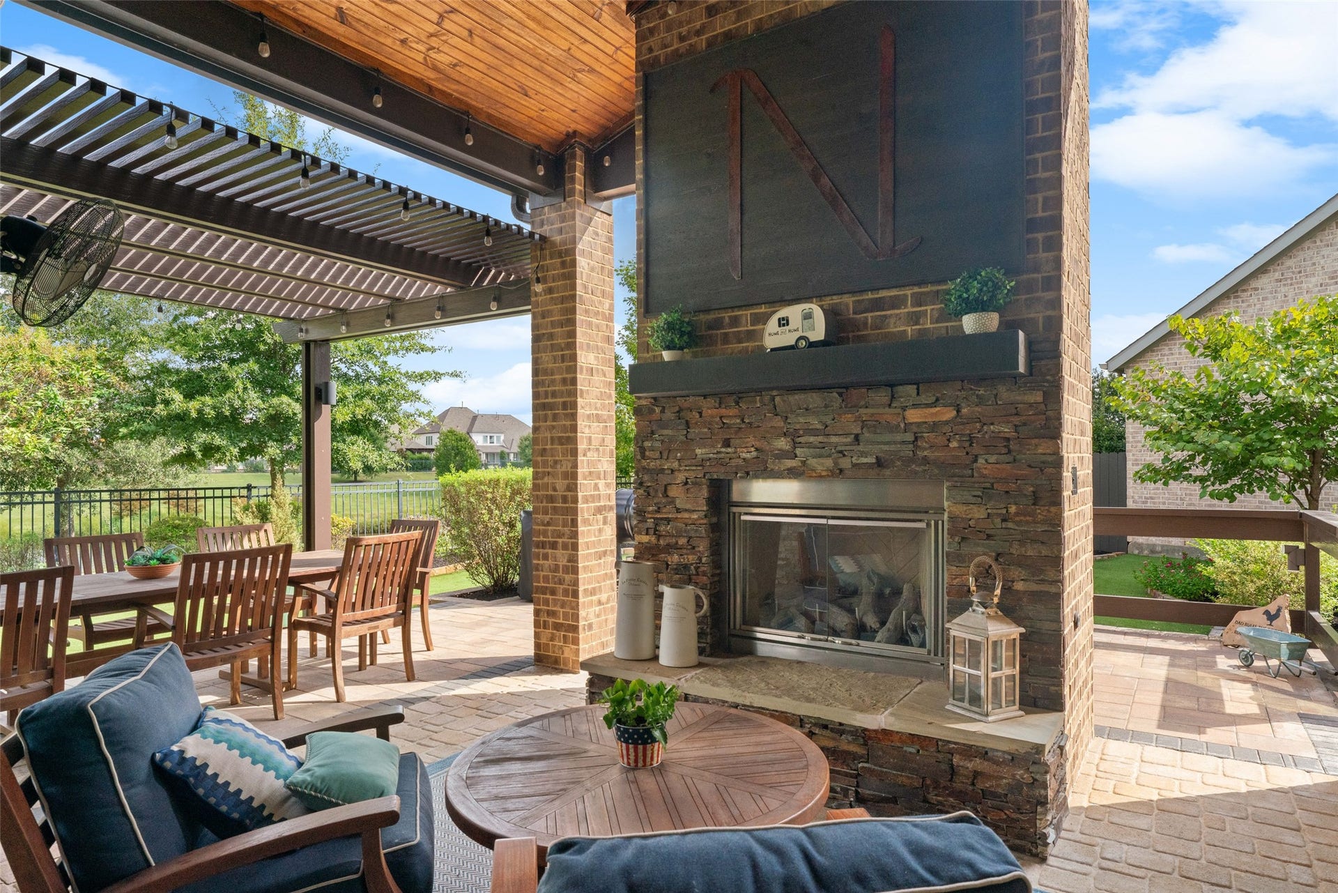 23926 Arcola Glen Lane, Katy, TX 77493 Custom outdoor gas fireplace featuring a stone and brick chimney with a raised hearth, wood mantle, and an integrated TV mount with cover. Located under a covered lanai adjacent to a two-tier metal pergola, creating a comfortable space for outdoor dining and relaxation.