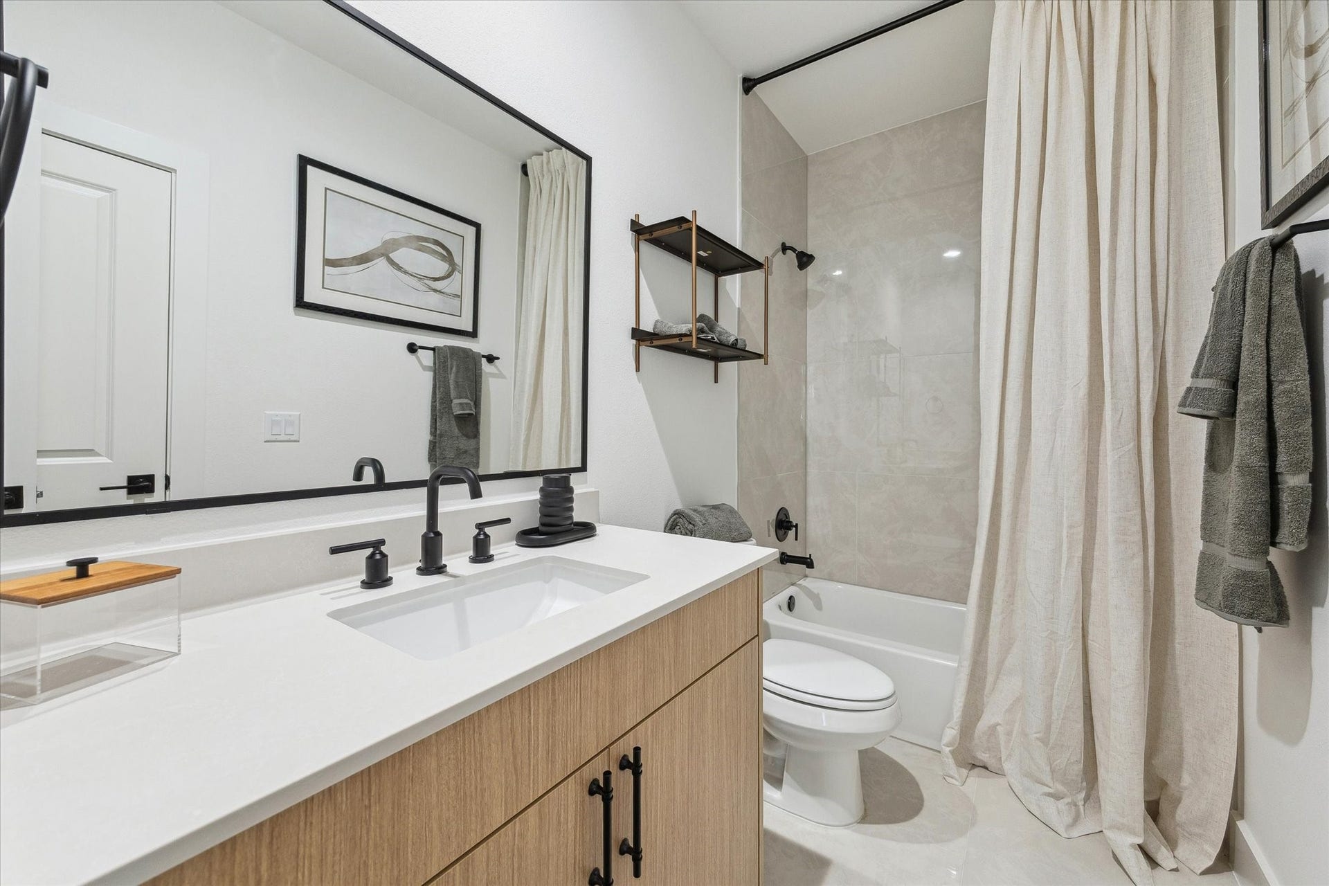 Sleek bathroom featuring a modern vanity with black fixtures, a spacious mirror, and a bathtub with a chic tile surround. Neutral tones and tasteful decor create a calming atmosphere.