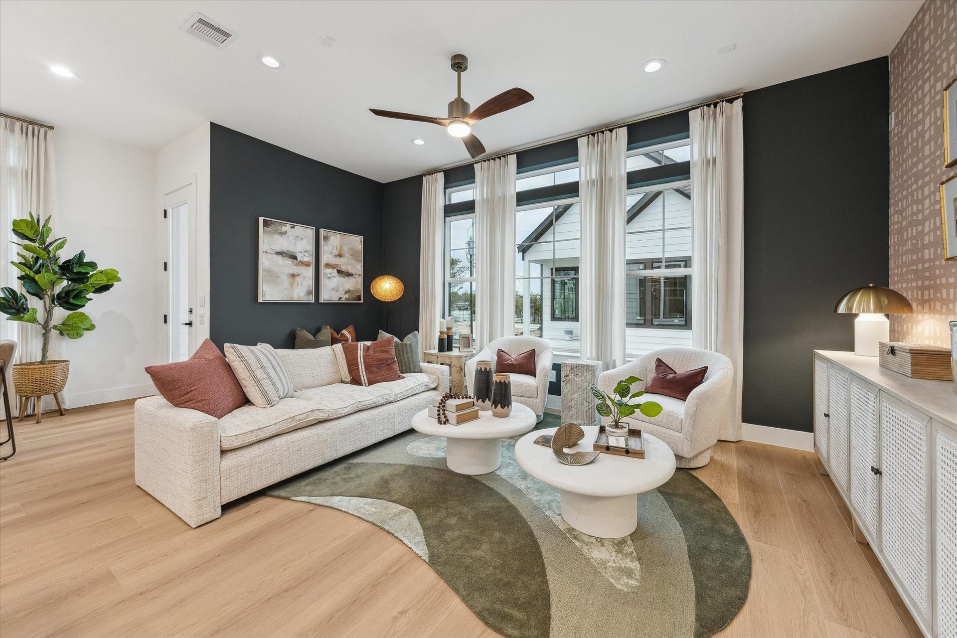 Elegant living room with sleek wood flooring, a cozy sectional sofa, and large windows offering abundant natural light. Modern decor and a stylish ceiling fan add a touch of sophistication.