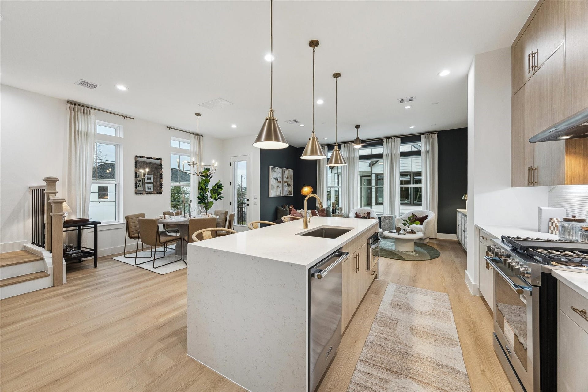 Sleek open-concept kitchen and living area featuring a spacious island with modern pendant lighting, stainless steel appliances, and elegant dining space. Natural light pours in through large windows, highlighting the contemporary design and warm wood flooring.