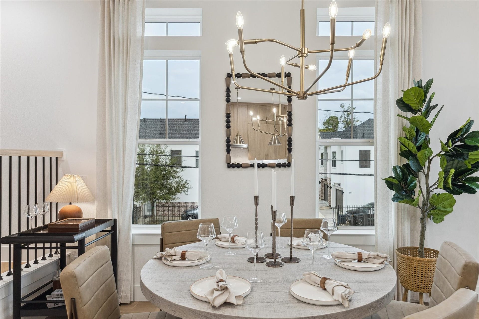 Elegant dining area featuring a round table set for four, under a striking modern chandelier. Large windows provide ample natural light, complemented by tasteful decor and a lush potted plant for a welcoming ambiance.