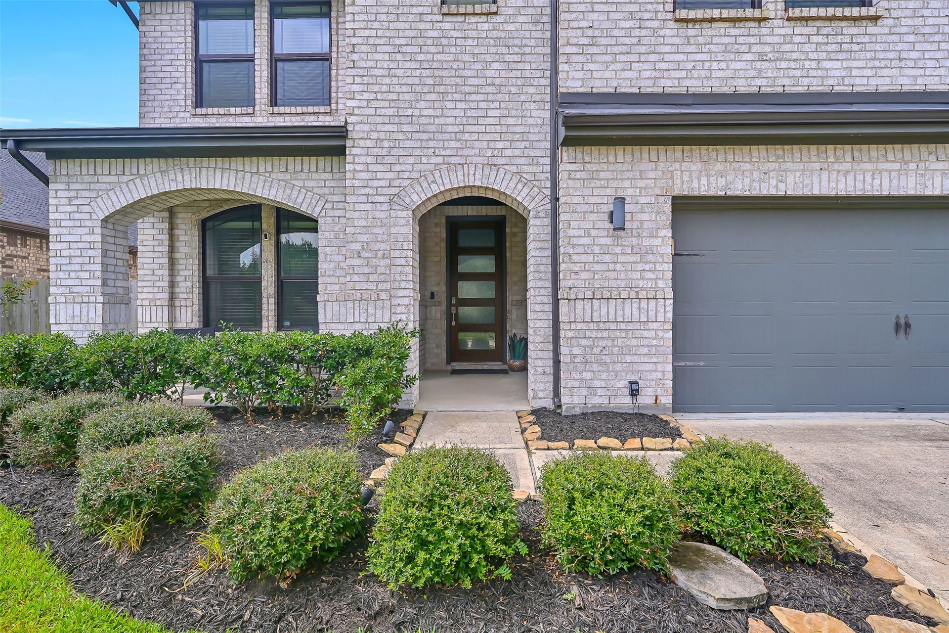 1570 Mustang Trail, Friendswood, TX 77546 A closer look at the inviting front elevation showcases arched architectural details, lush landscaping, a covered front porch, and a contemporary wood-and-glass entry door that offers a stylish introduction to the home.
