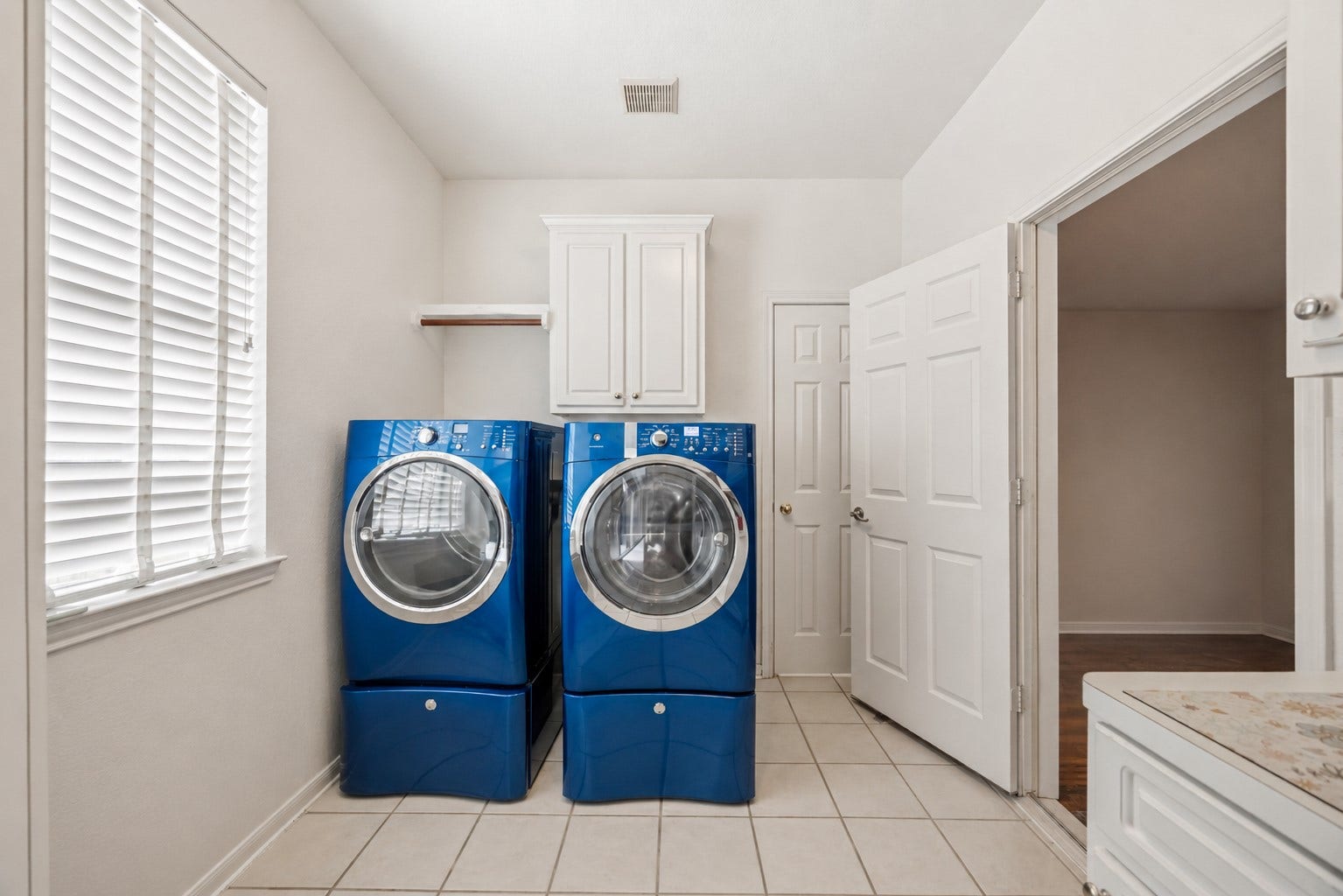 7610 Guinevere Drive, Sugar Land, TX 77479 The dedicated laundry room includes white upper cabinetry, a hanging rod, tile floors, and a side window with horizontal blinds.