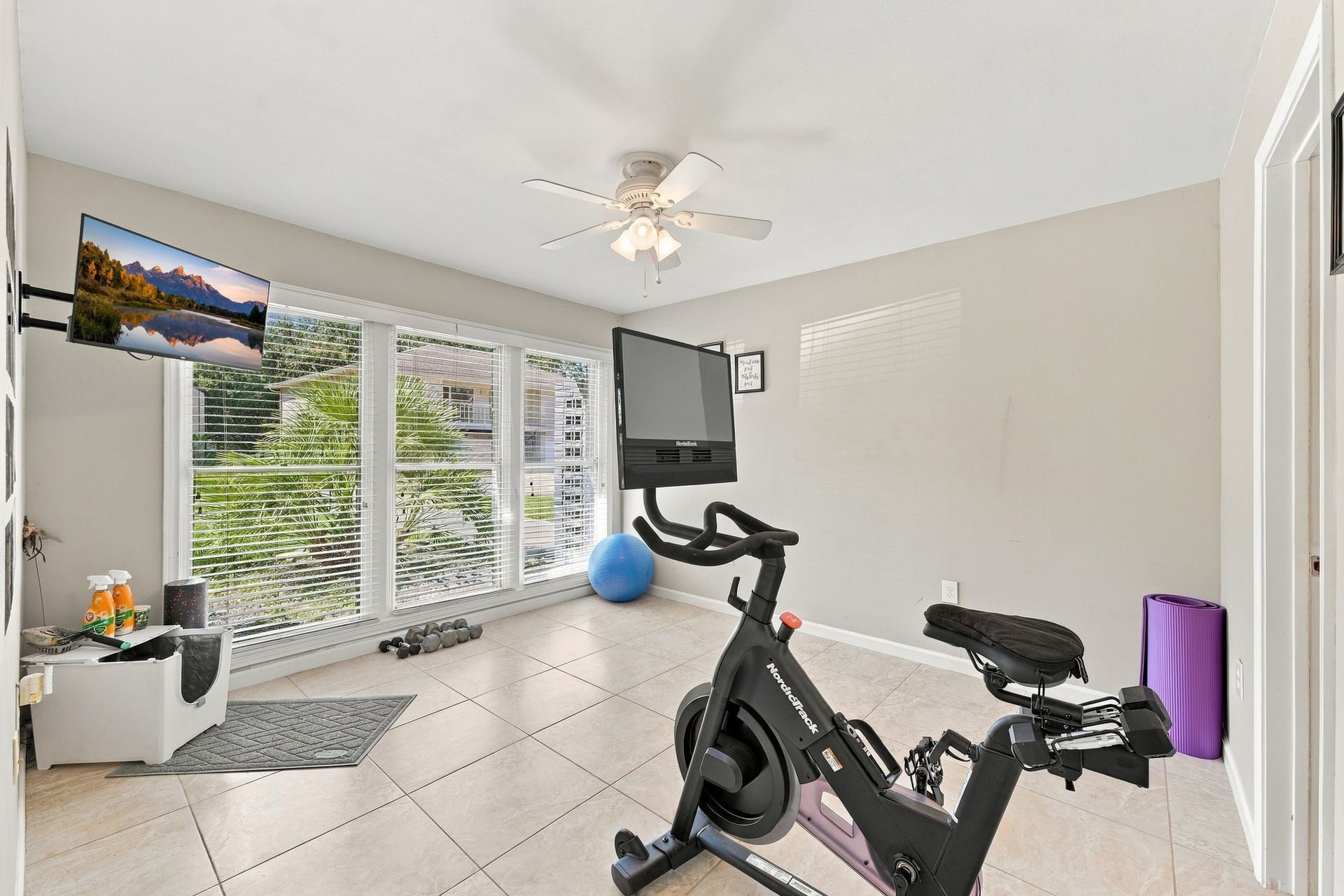 10 Harbor Lane, Kemah, TX 77565 The third bedroom is currently being used as a exercise room with large windows, tiled flooring, and a ceiling fan, offering a perfect space for workouts with a stationary bike and mounted TV.