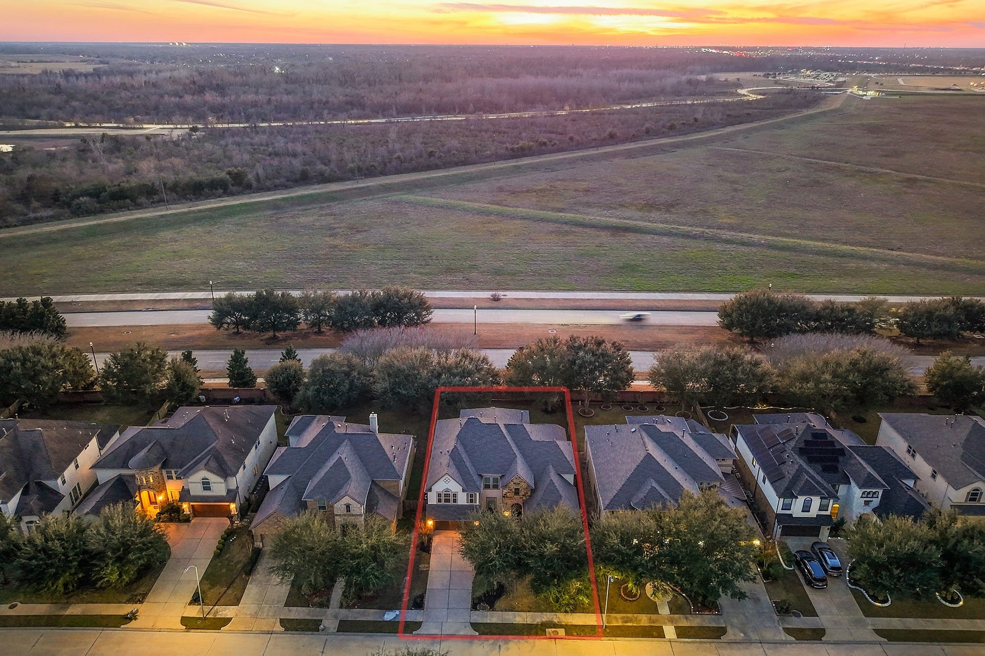 3102 Barrons Way, Sugar Land, TX 77479 This aerial photo showcases a row of suburban homes with well-maintained lawns and driveways, set against the backdrop of an open field, offering a serene and spacious environment. The sunset in the distance adds a warm, inviting glow to the neighborhood.