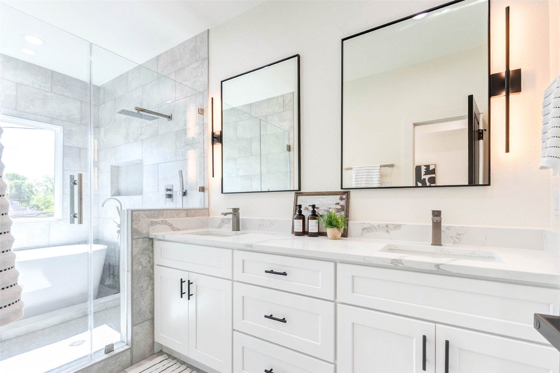 The dual-sink vanity is topped with marble-style quartz and accented by minimalist black hardware and vertical sconce lighting.