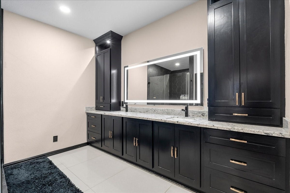 7202 Pine Drive W, Alvin, TX 77511 Elegant primary bathroom featuring sleek black cabinetry, a spacious dual-sink vanity with a granite countertop, and a modern backlit mirror.
