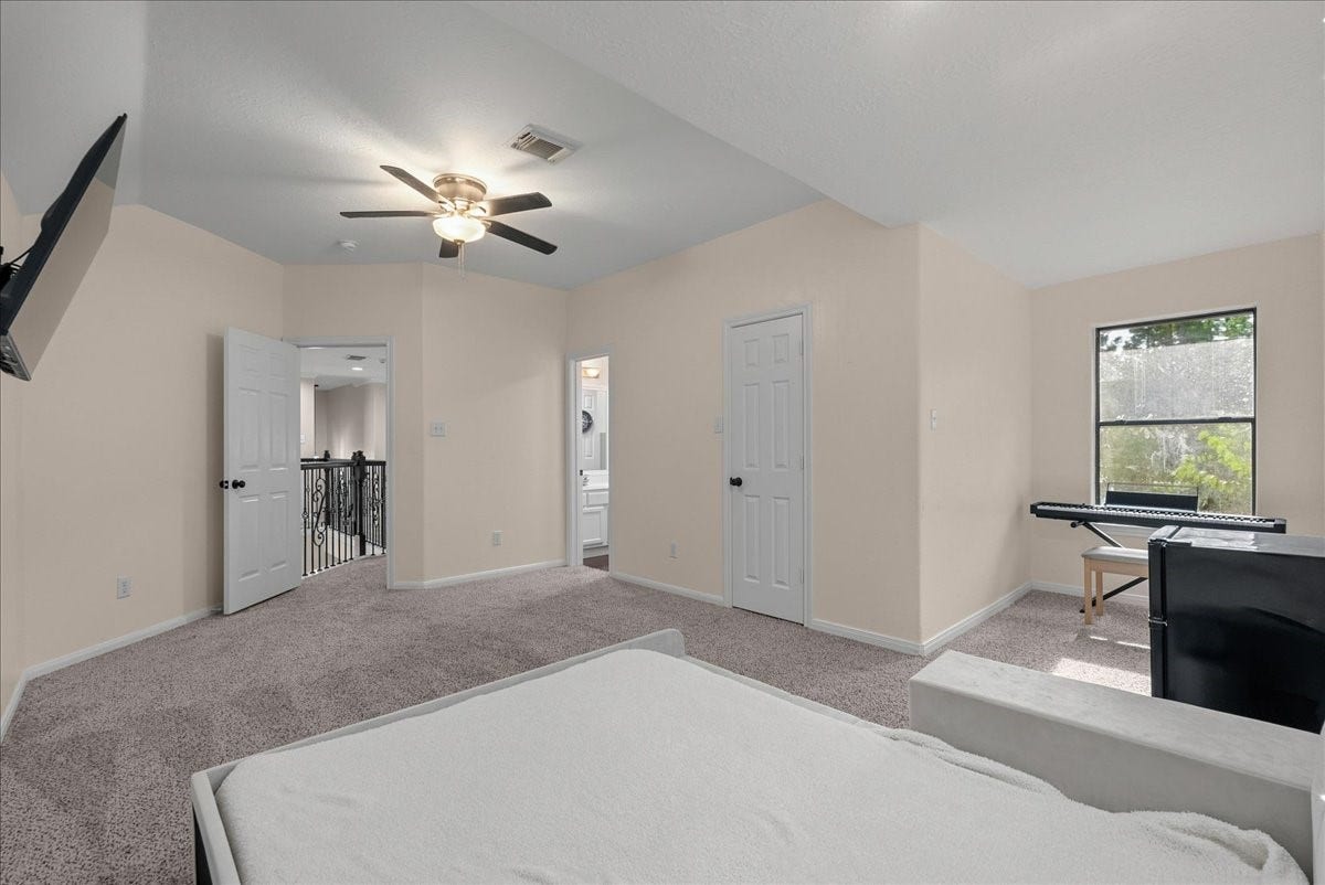 7202 Pine Drive W, Alvin, TX 77511 Spacious bedroom with plush carpeting, featuring a ceiling fan, large window for natural light, and convenient access to an en-suite bathroom and hallway.