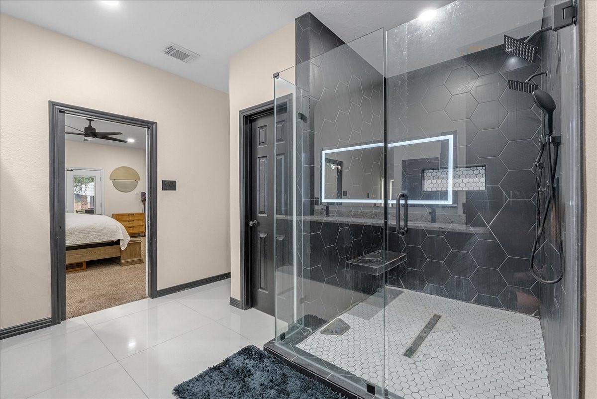 7202 Pine Drive W, Alvin, TX 77511 Luxurious primary bathroom featuring a spacious glass-enclosed shower with sleek hexagonal tiles and modern fixtures, complemented by bright, polished flooring. A glimpse into the cozy adjoining bedroom adds to the inviting atmosphere.