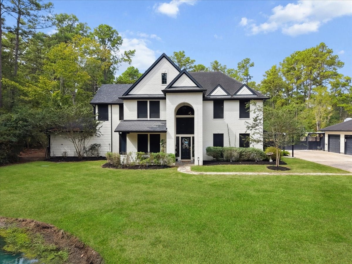 7202 Pine Drive W, Alvin, TX 77511 Elegant two-story home with a striking black and white faade, surrounded by lush greenery and nestled in a serene, wooded setting.