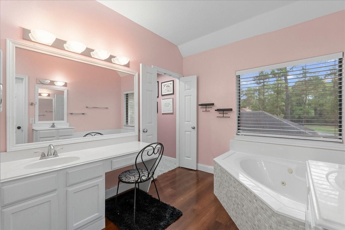 7202 Pine Drive W, Alvin, TX 77511 Charming upstairs primary bathroom featuring a spacious vanity with ample lighting, a cozy makeup area, elegant wood like flooring, and a relaxing corner bathtub by a bright window with scenic views.