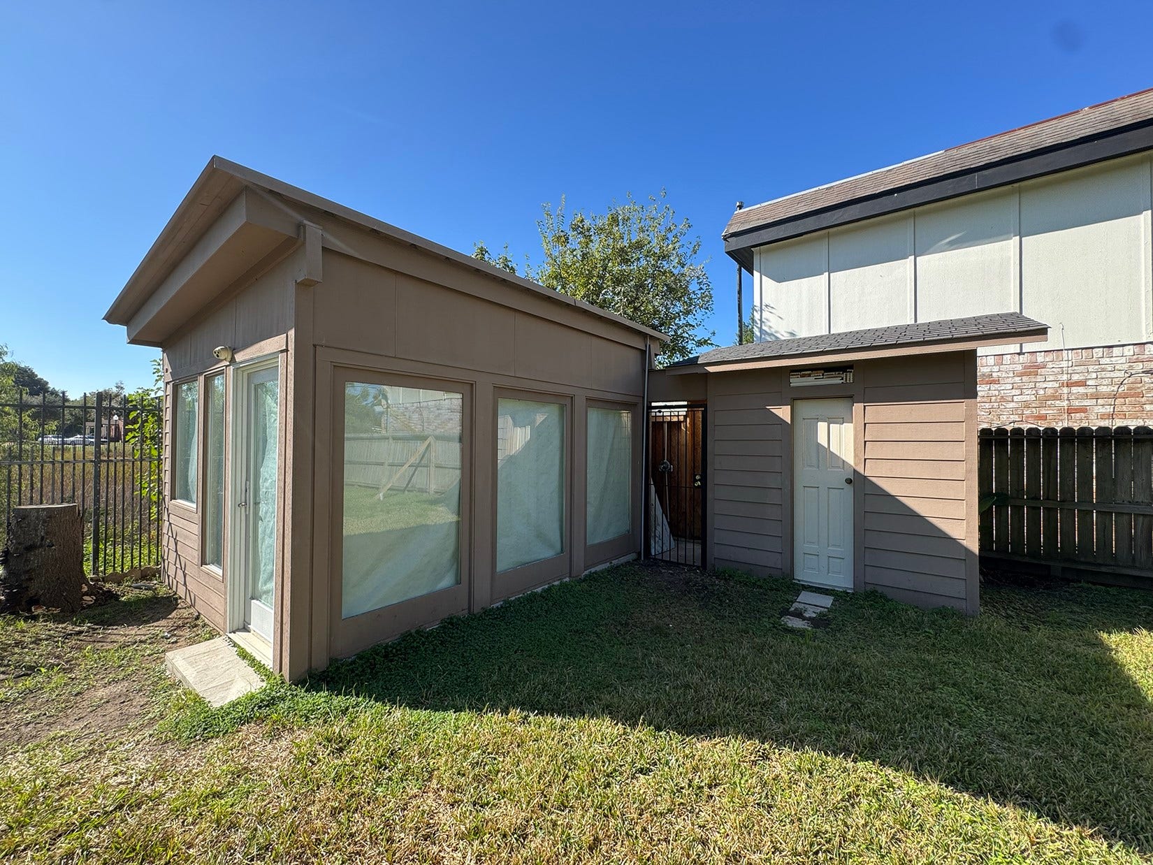 This photo shows a small backyard structure with large windows, adjacent to another building. It has a fenced yard with grass and appears to receive ample sunlight. Ideal for extra storage space.