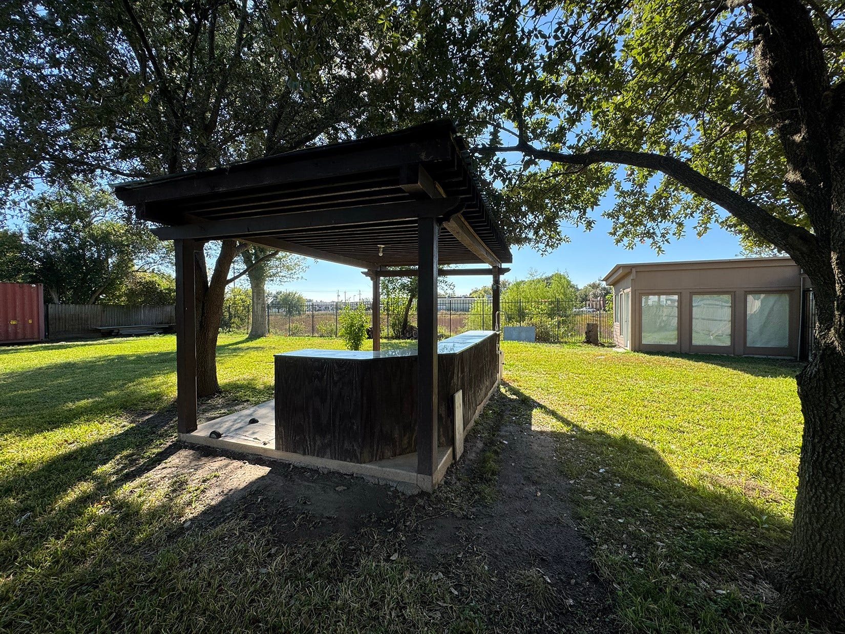 Spacious backyard featuring a cozy wooden gazebo with a countertop, perfect for outdoor entertaining, surrounded by mature trees and a well-maintained lawn. A separate building with large windows complements the space, offering potential for extra storage.