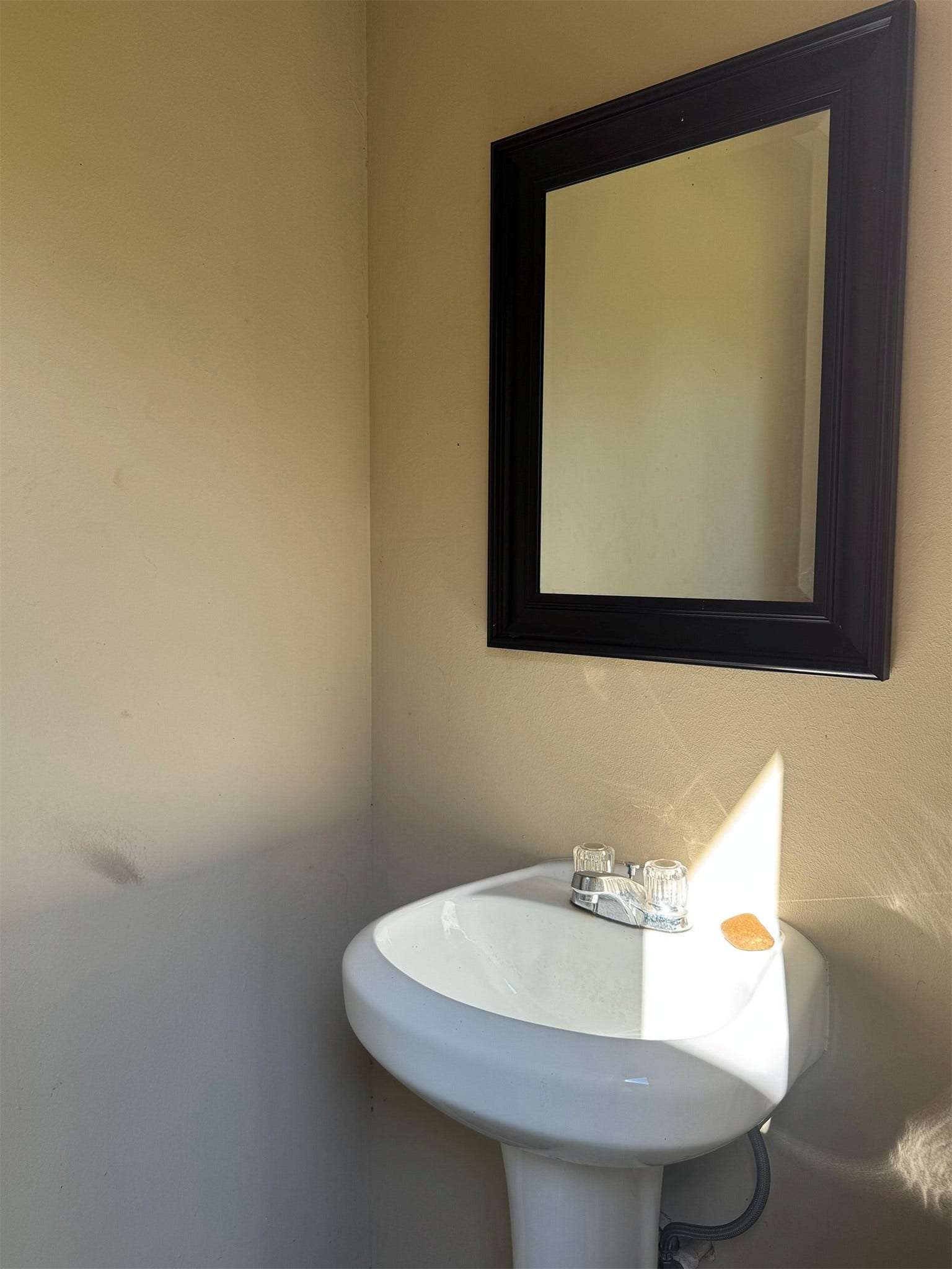 Well-lit bathroom corner with a white pedestal sink and a wall-mounted mirror. The neutral wall color adds to the clean and minimalistic feel.