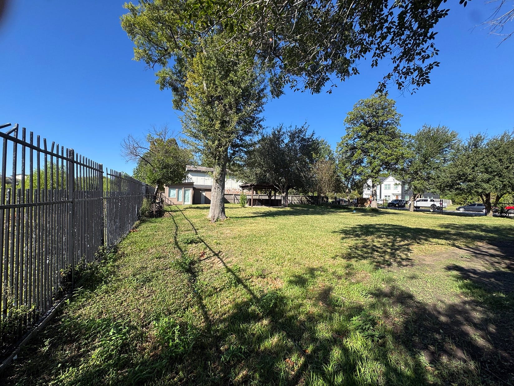 Spacious, grassy lot with mature trees, a secure metal fence, and a clear blue sky. Ideal for building or landscaping, offering privacy and tranquility.