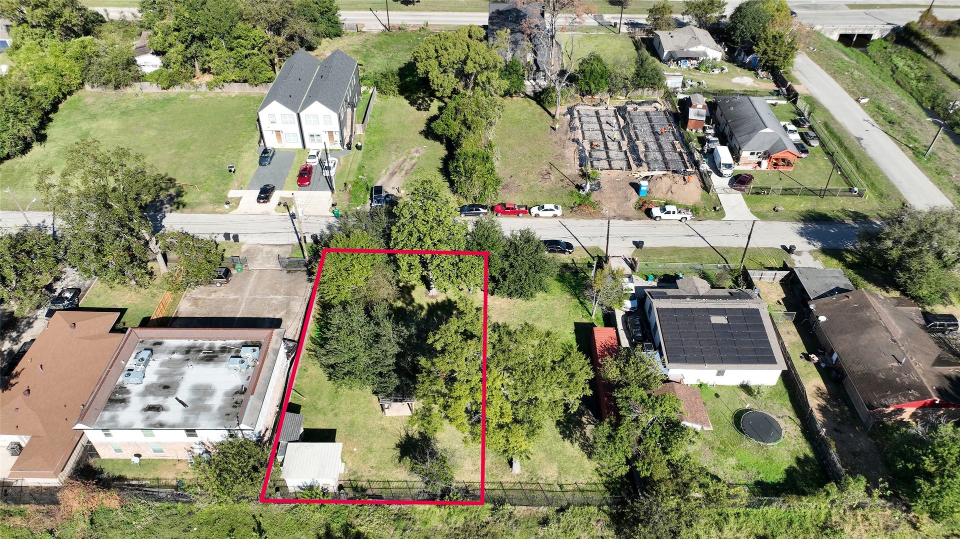 This aerial photo shows a residential lot outlined in red, located in a suburban neighborhood. The lot is surrounded by a mix of existing homes, some with solar panels, and nearby open spaces. It's a green, tree-lined area with easy access to roads. Perfect for building new homes.