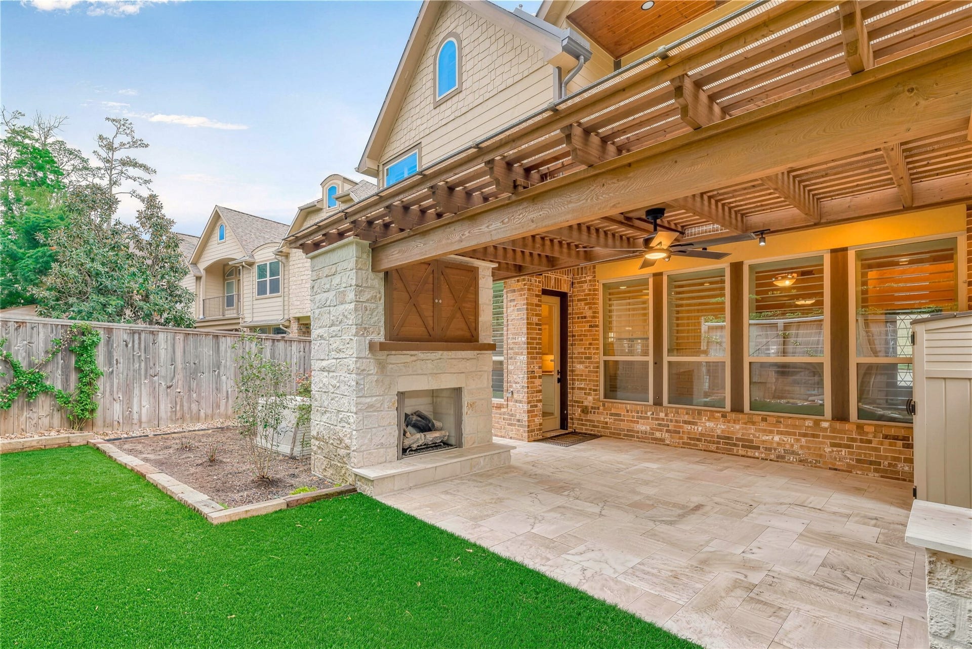 19 Herons Flight Place, Spring, TX 77389 Charming outdoor living space featuring a cozy stone fireplace under a pergola, complemented by lush artificial turf and elegant tile flooring for seamless indoor-outdoor entertaining.