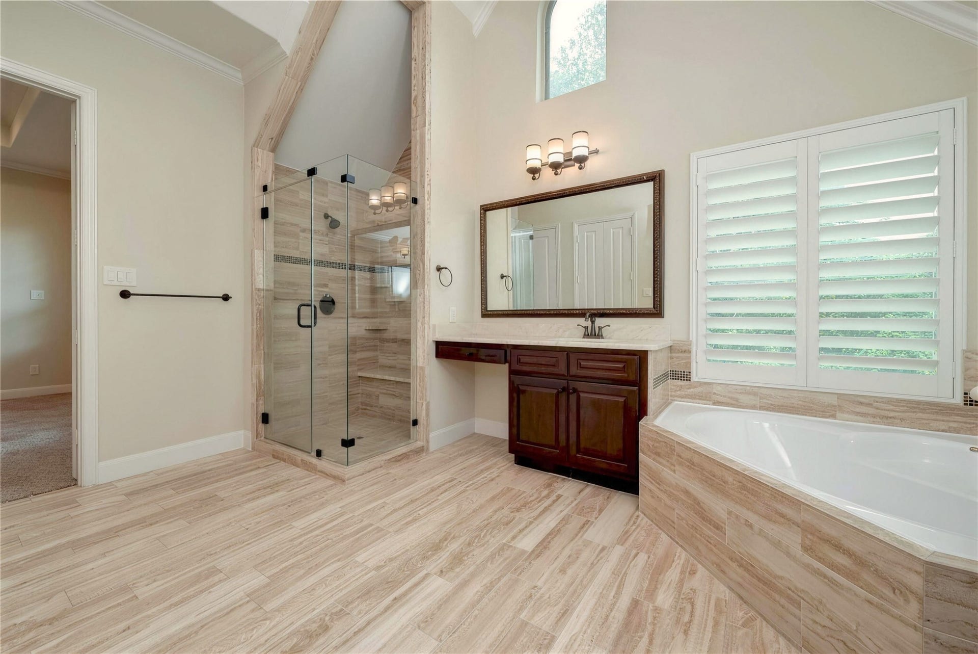 19 Herons Flight Place, Spring, TX 77389 Elegant bathroom featuring a spacious glass-enclosed shower, luxurious soaking tub, and rich wood vanity. Natural light pours in through large windows with plantation shutters, highlighting the serene atmosphere.