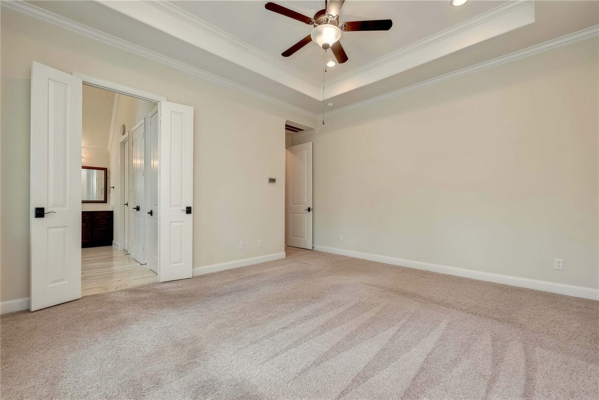 19 Herons Flight Place, Spring, TX 77389 Spacious bedroom featuring plush carpeting, elegant tray ceiling with recessed lighting, and a stylish ceiling fan. Double doors open to an adjoining bathroom, adding convenience and luxury.