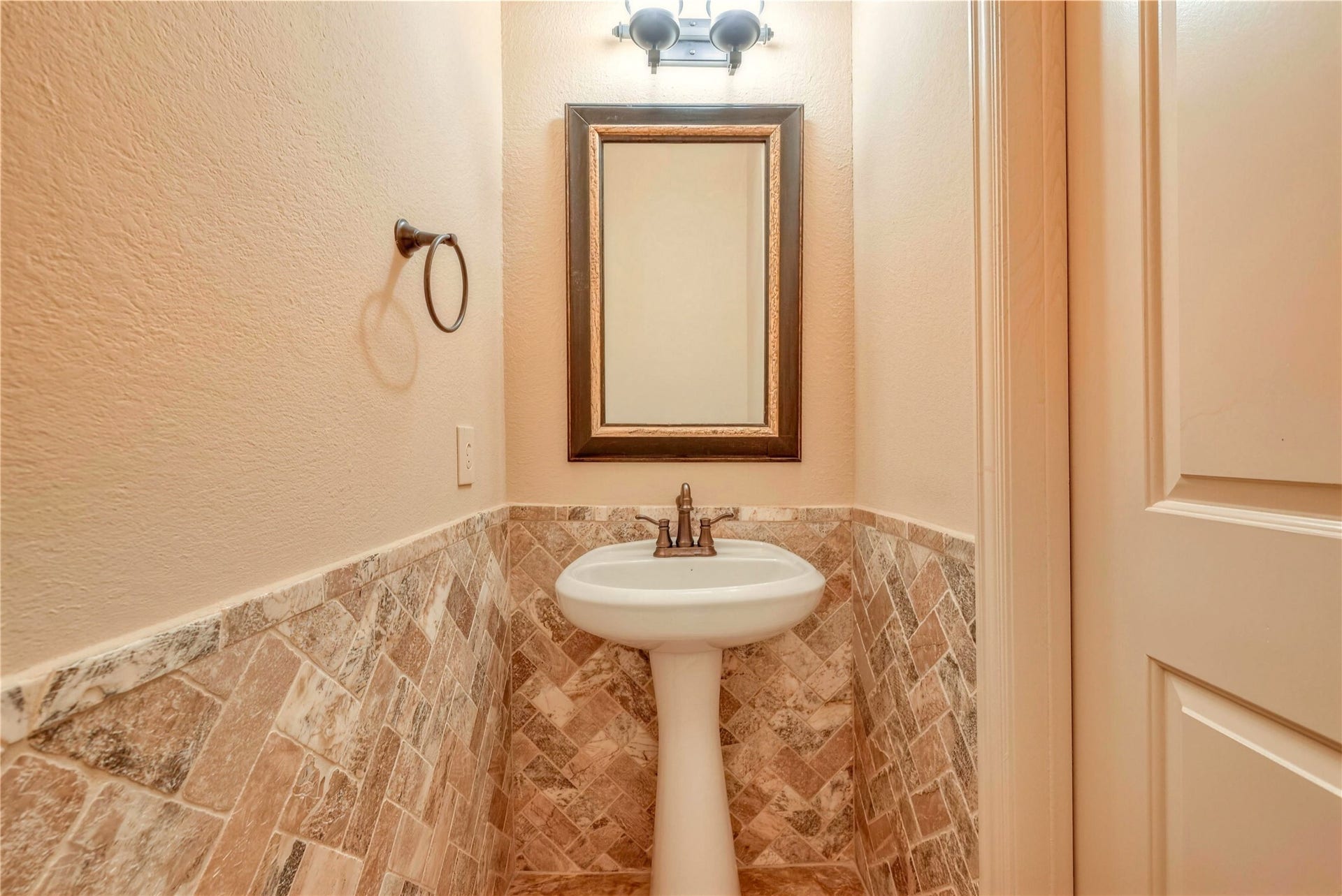 19 Herons Flight Place, Spring, TX 77389 Charming powder room featuring elegant stone tile walls, a classic pedestal sink, and a stylish framed mirror under soft lighting.
