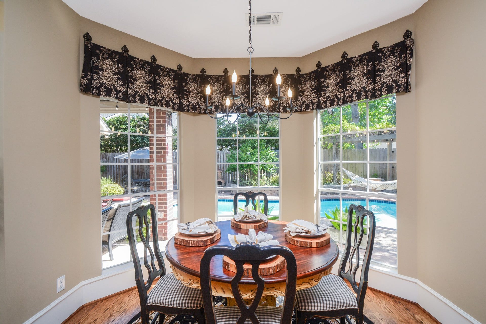 A bright breakfast nook features bay windows overlooking the backyard pool and hardwood flooring beneath a decorative chandelier.