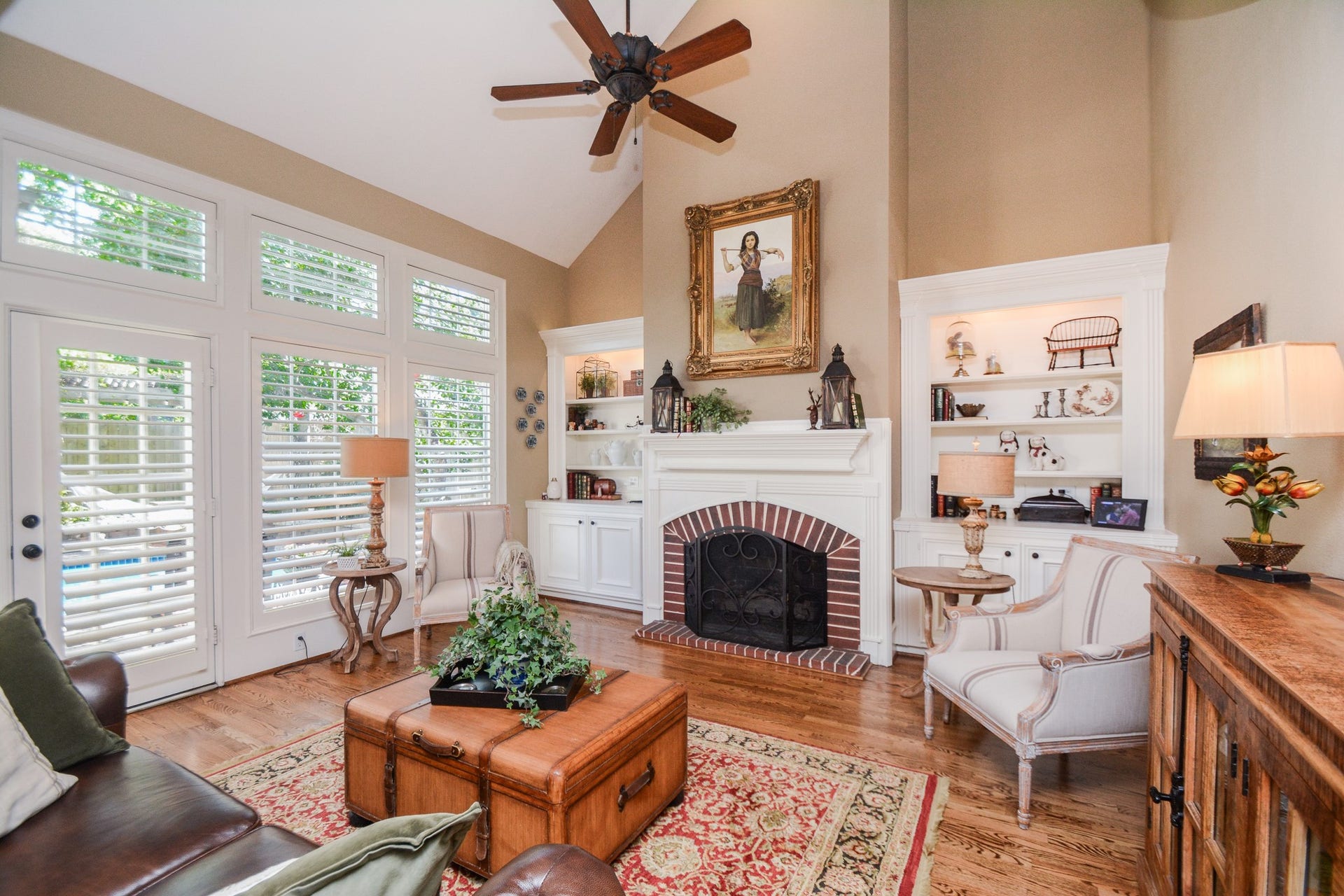 Tall windows with plantation shutters fill the family room with natural light beside the fireplace and built-in display shelves.