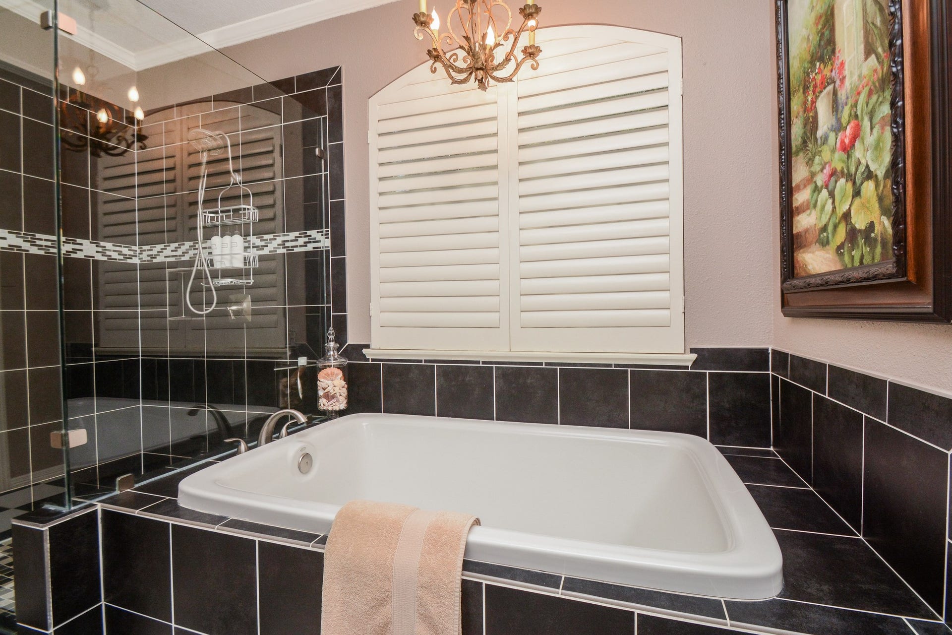 Unwind in the primary bathrooms soaking tub beneath a decorative chandelier, surrounded by striking black tile and plantation shutters.
