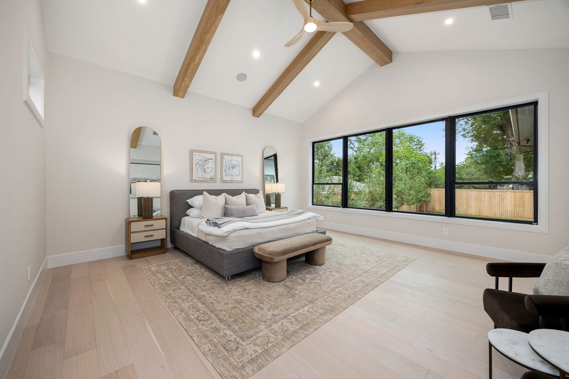 14106 Heatherfield Drive, Houston, TX 77079 A serene and spacious primary bedroom designed for comfort and relaxation, featuring wide-plank flooring and a soft, neutral palette. Large picture windows flood the room with natural light while framing views of the outdoors. The highlight of this space is the vaulted ceiling with exposed wood beams, adding architectural character and warmth while creating an open, airy retreat that feels both luxurious and inviting.