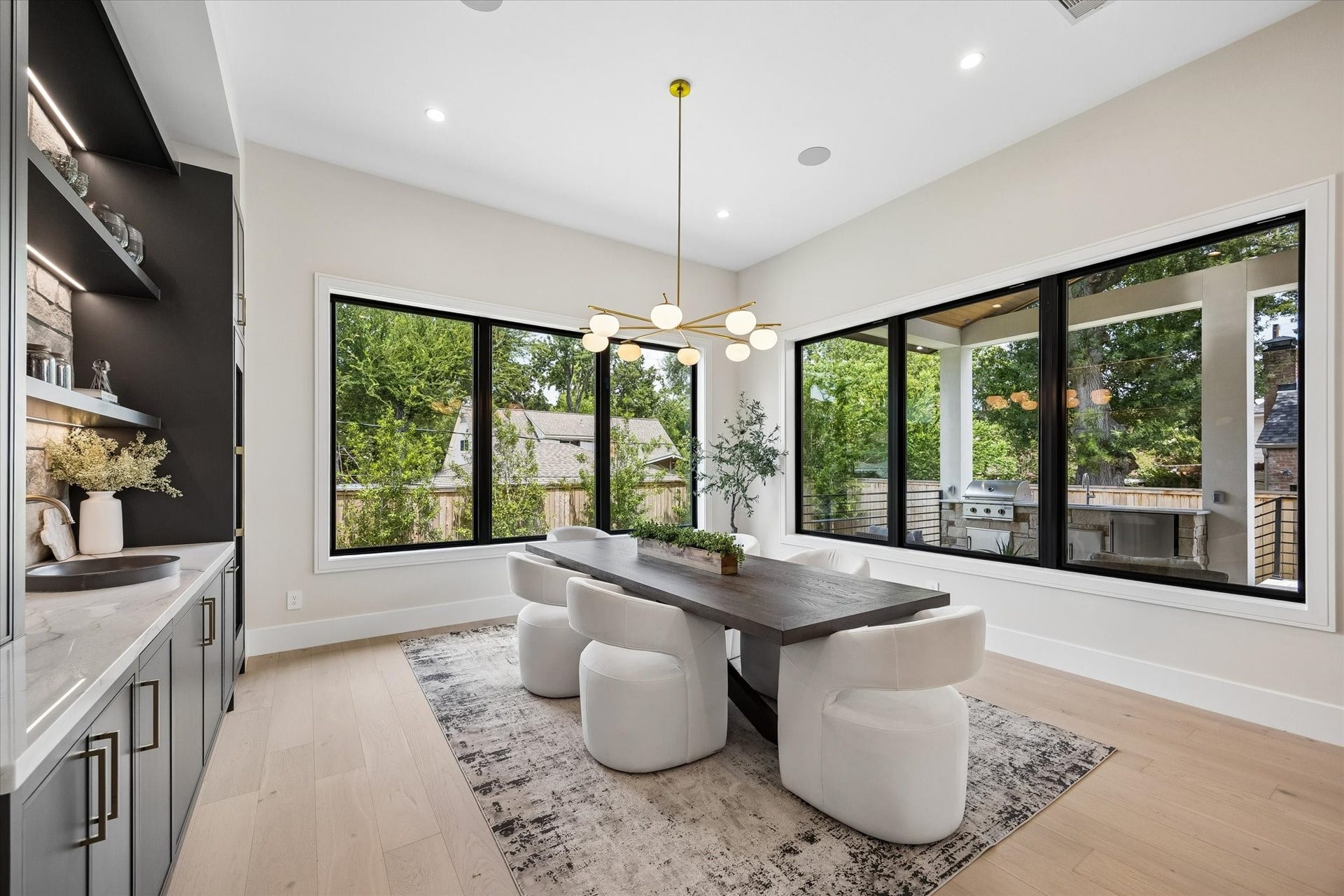 14106 Heatherfield Drive, Houston, TX 77079 Walls of black-framed windows turn this dining area into a bright, garden-connected retreat. Custom cabinetry with integrated lighting, stone detailing and a sleek beverage station adds both beauty and function, while the sculptural chandelier brings a refined focal point overhead. The room flows effortlessly toward the covered outdoor living area, creating a natural connection between indoor entertaining and relaxed open-air dining.